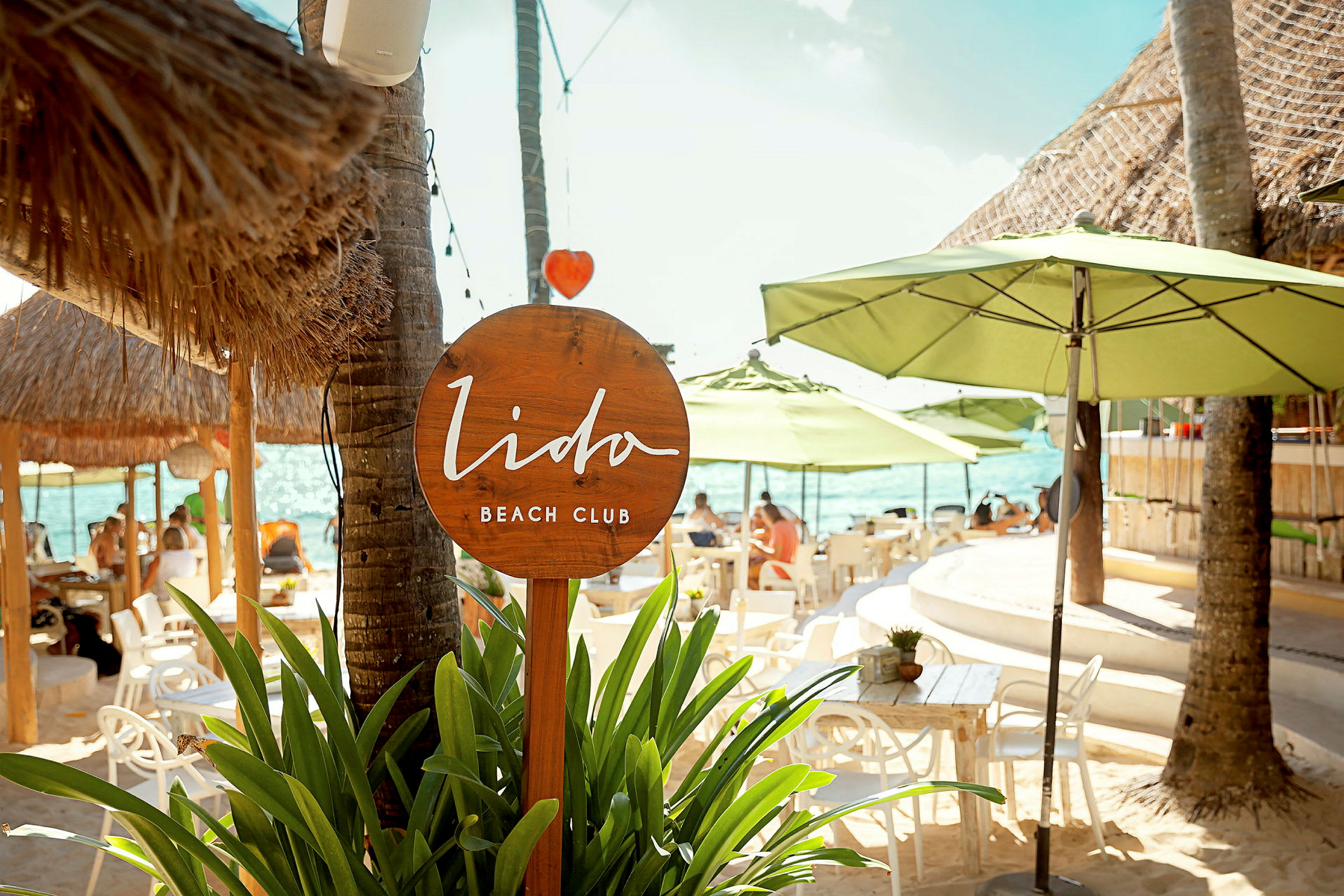 En skylt för Lido Beach Club omgiven av gröna växter och parasoller på en strand. (AI-genererad text)