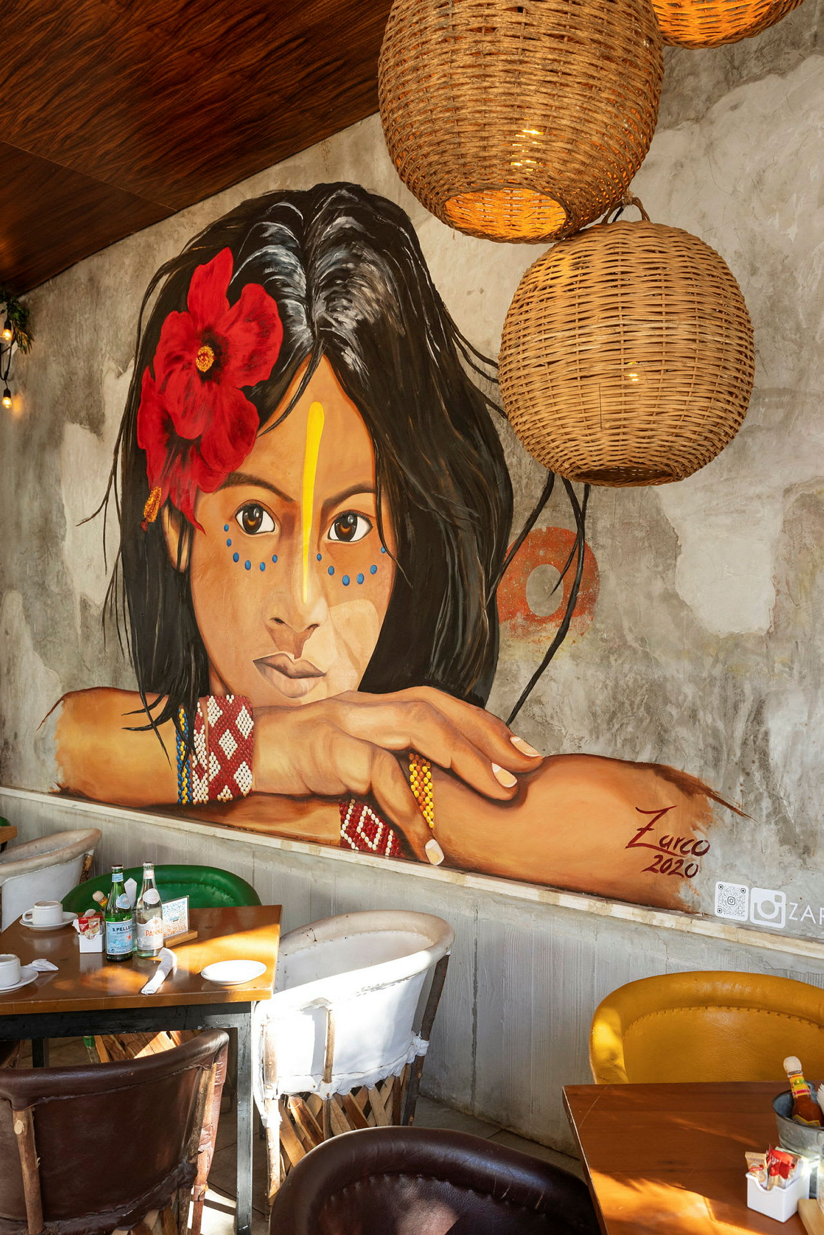 En väggmålning av en ung kvinna med en röd blomma i håret och färgglada mönster, omgiven av inredning i en restaurang. (AI-genererad text)