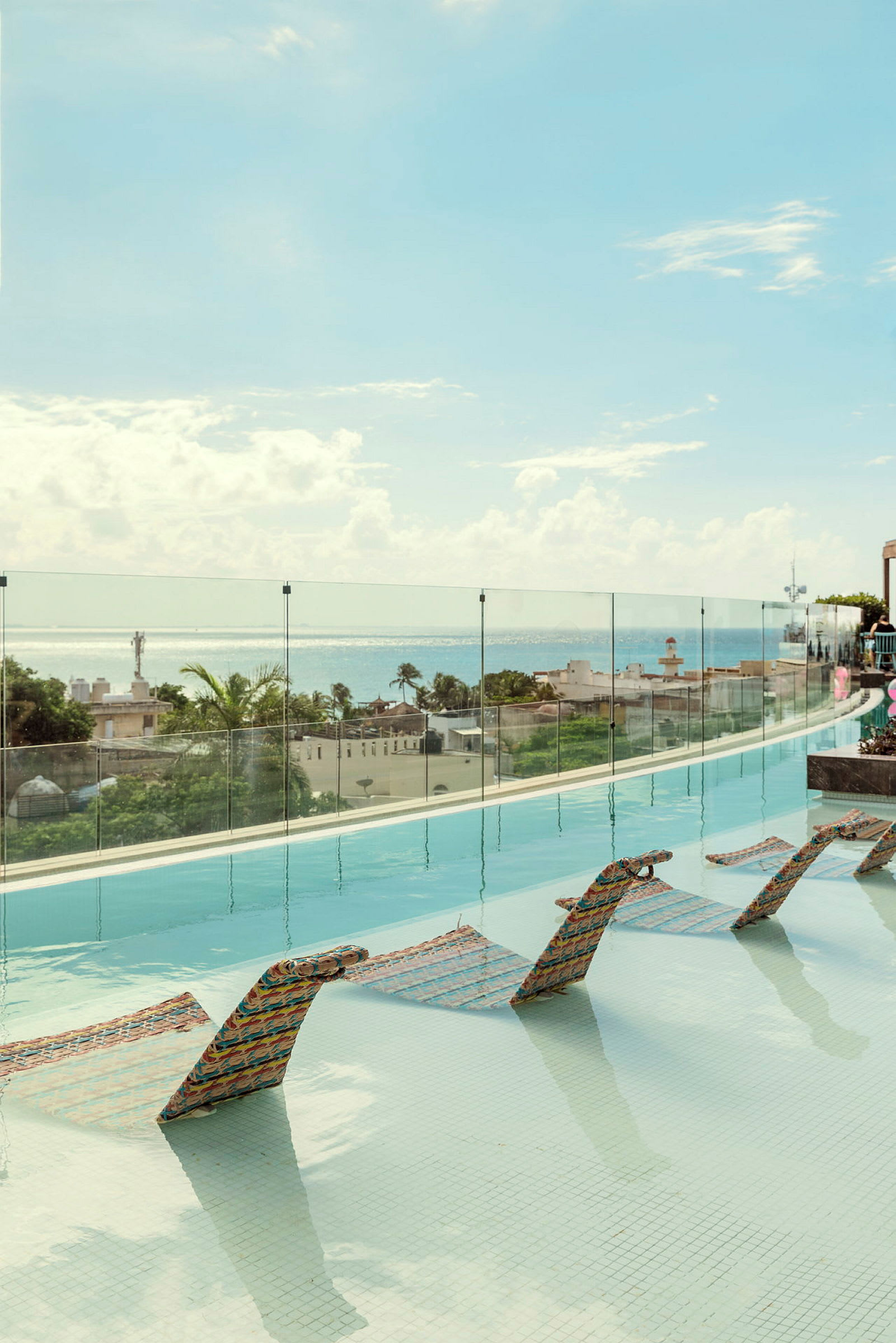 En infinitypool med solstolar i vattnet och utsikt över havet och hus i bakgrunden. (AI-genererad text)