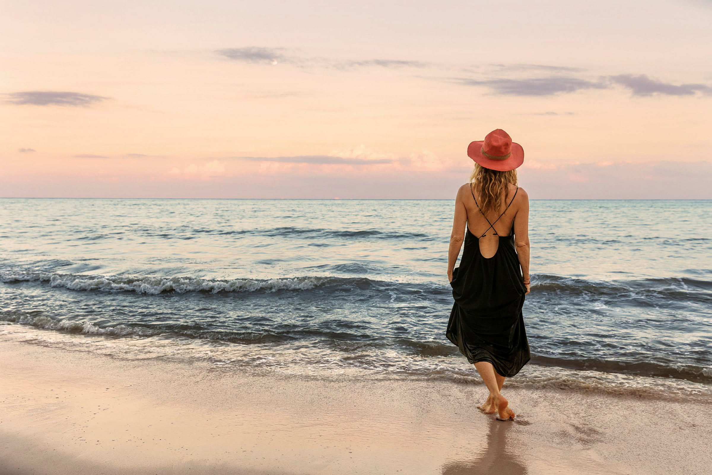 En person med en röd hatt och svart klänning går på en sandstrand vid havet under en pastellfärgad himmel. (AI-genererad text)