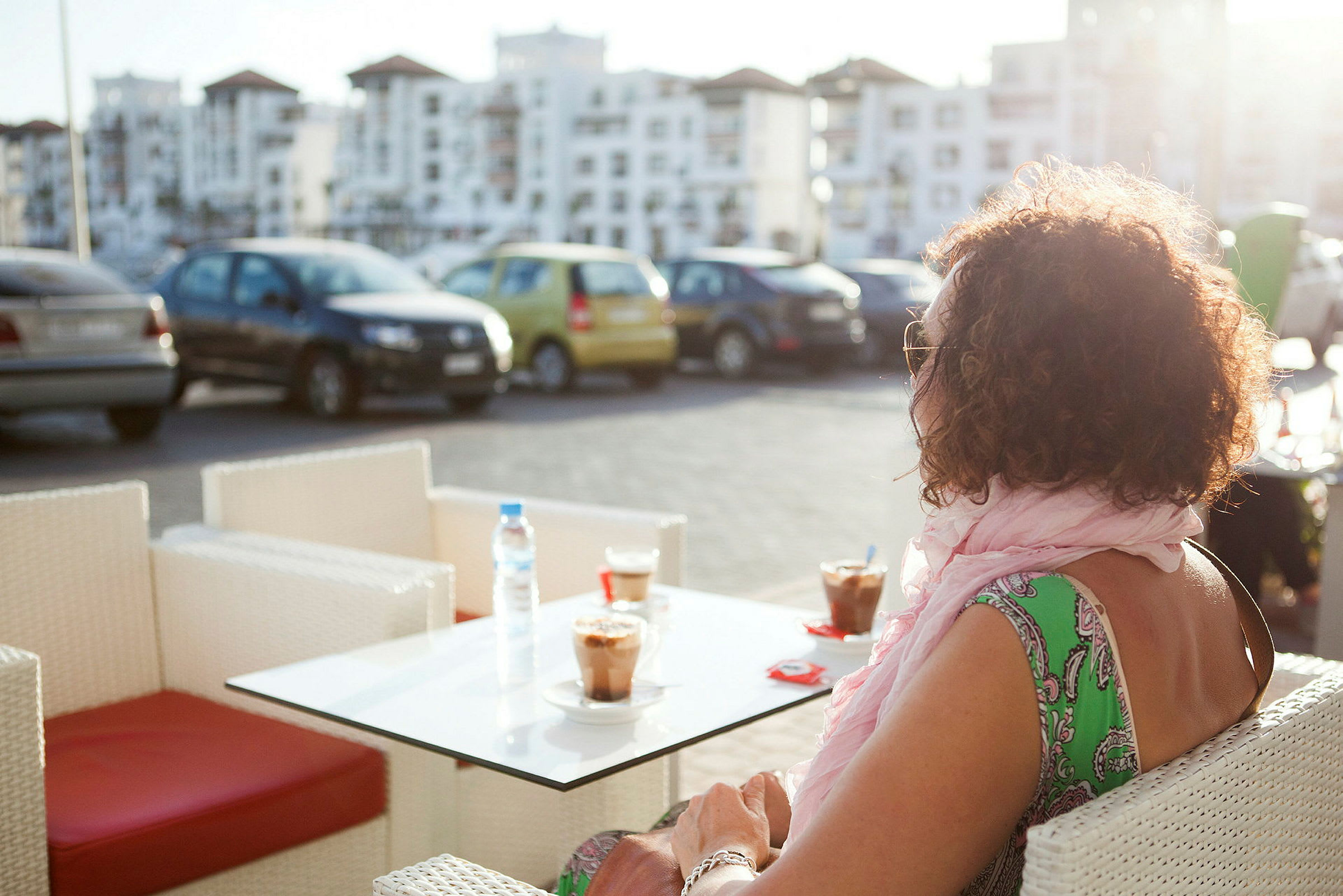 En person sitter vid ett bord utomhus med två kaffedrycker, i bakgrunden syns parkerade bilar och byggnader. (AI-genererad text)