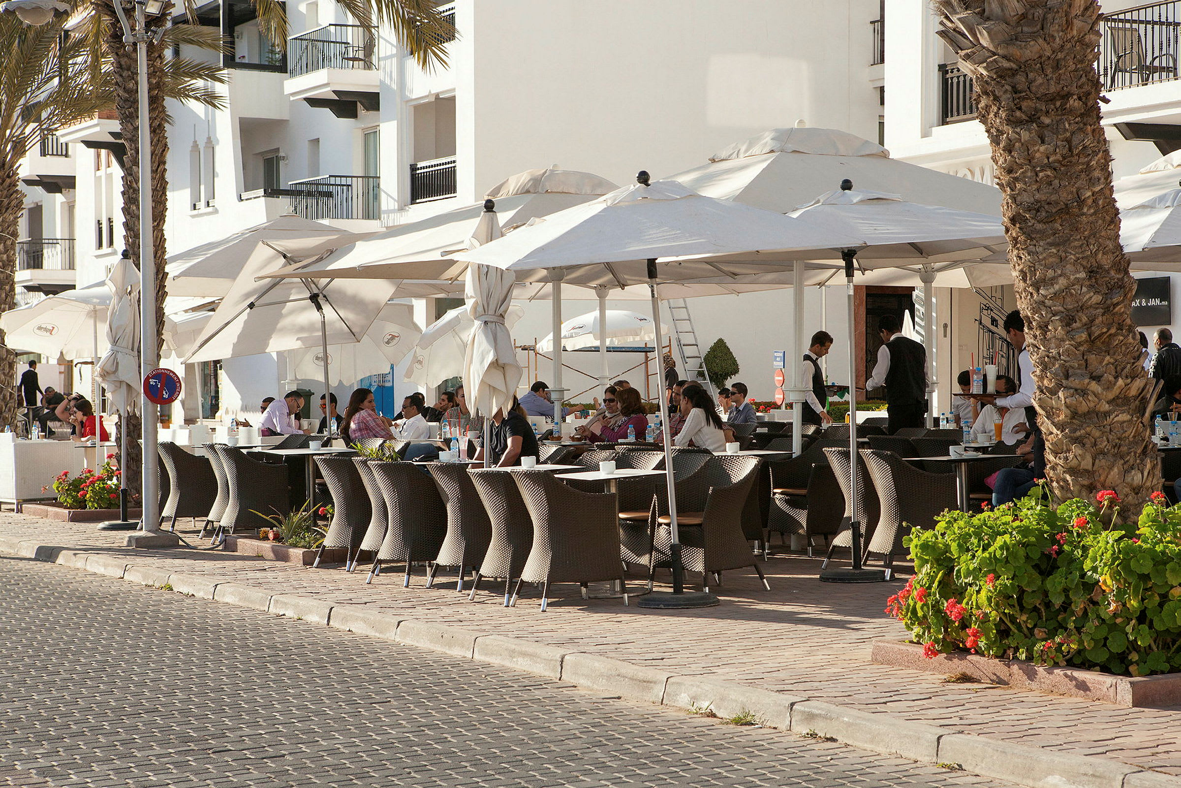 Utomhus restaurangområde med bord, stolar och parasoller vid ett stenbelagt gångstråk. (AI-genererad text)