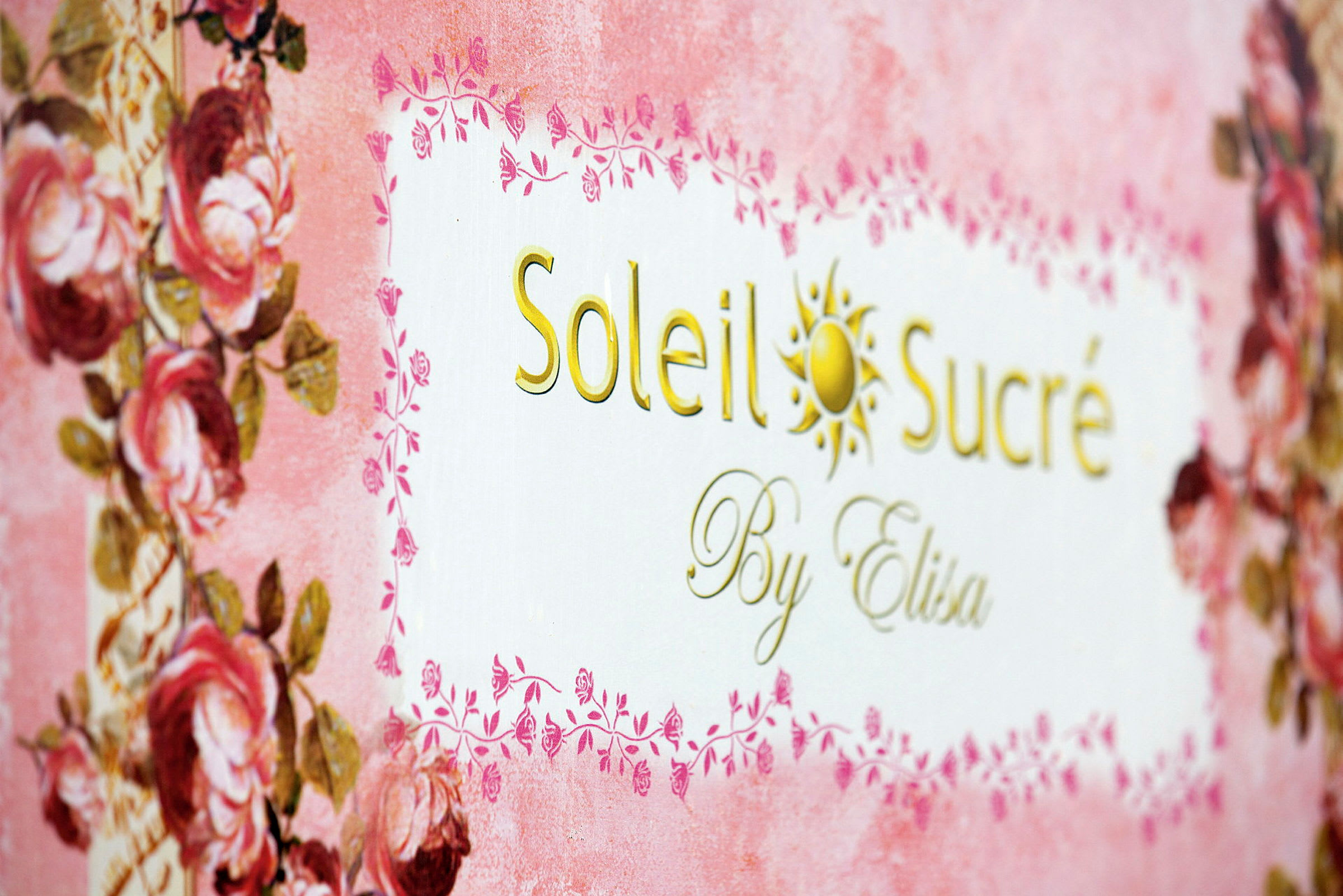 Texten 'Soleil Sucré By Elisa' på en dekorerad bakgrund med rosa och blomsterdetaljer. (AI-genererad text)