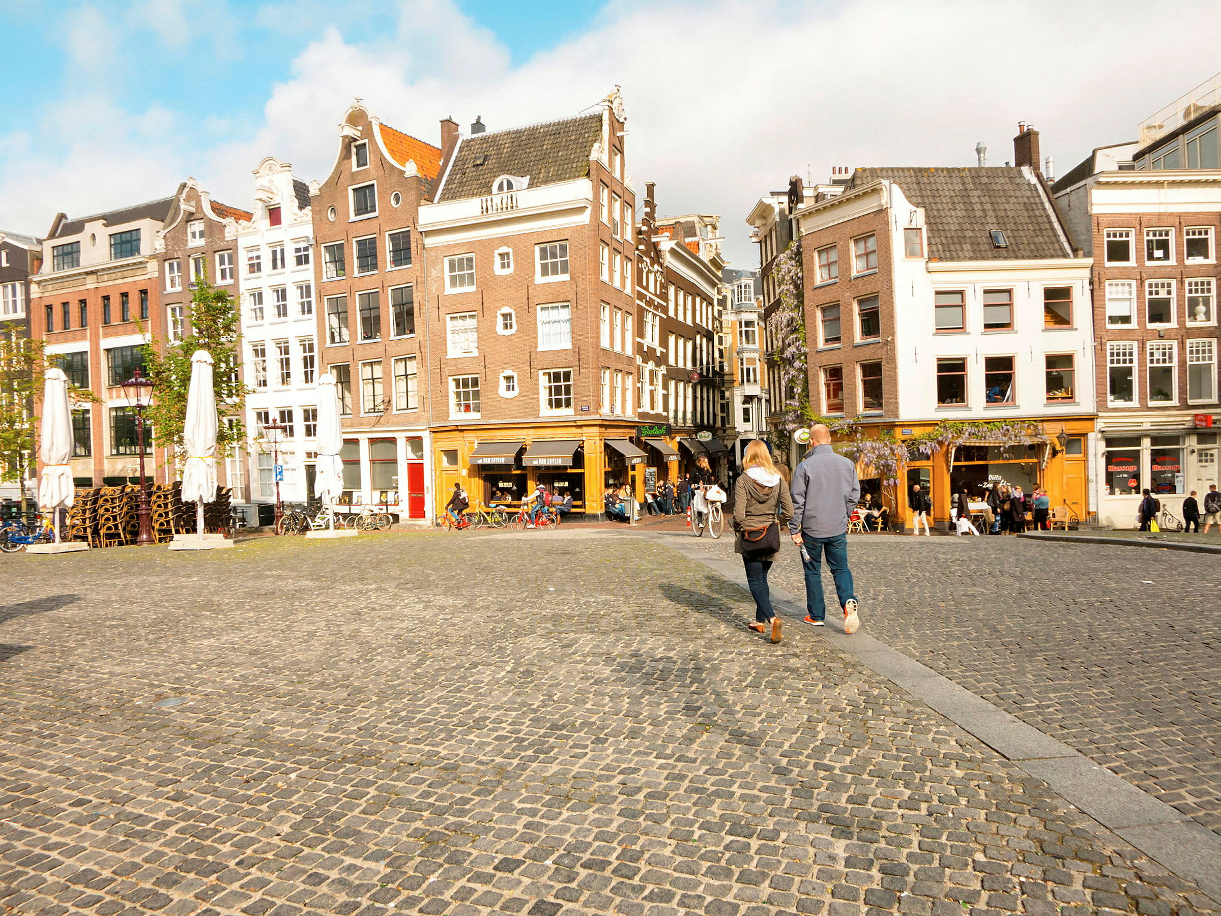 Bild av ett torg med kullersten och omgivande byggnader i klassisk europeisk stil. Två personer går över torget. (AI-genererad text)