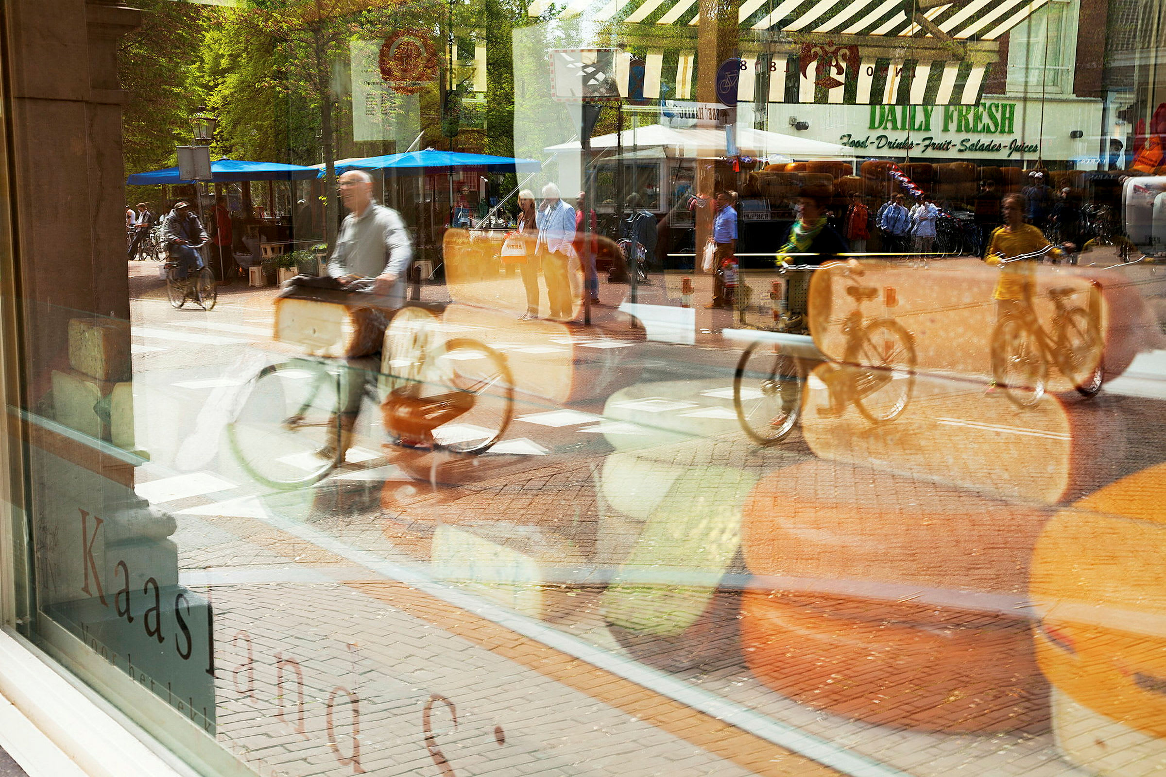 Ett skyltfönster med ost och en reflektion av en gata med cyklister och marknadsstånd syns. (AI-genererad text)