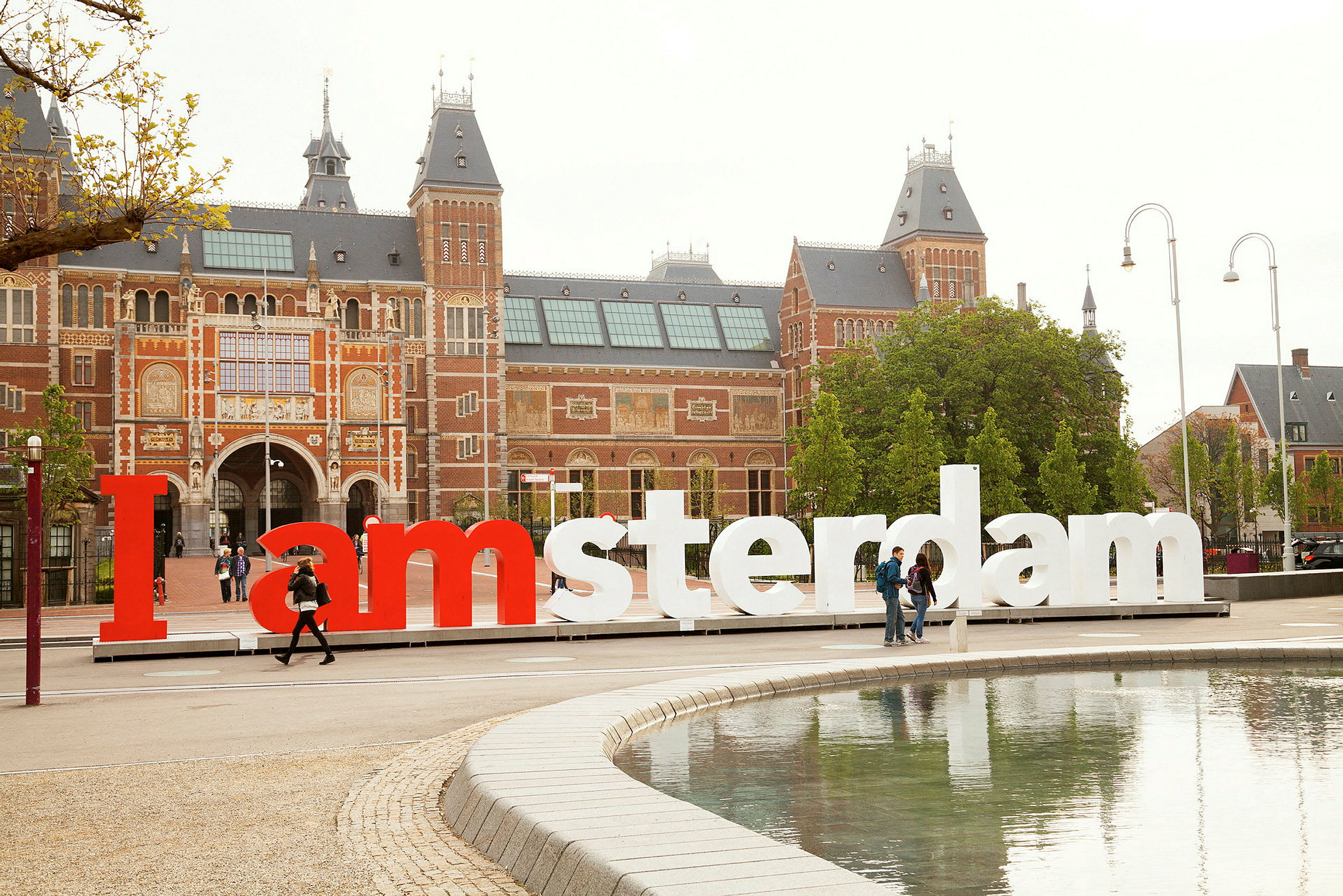 Stor byggnad med tegel och flera torn bakom en I amsterdam-skylt, vid vattnet. (AI-genererad text)