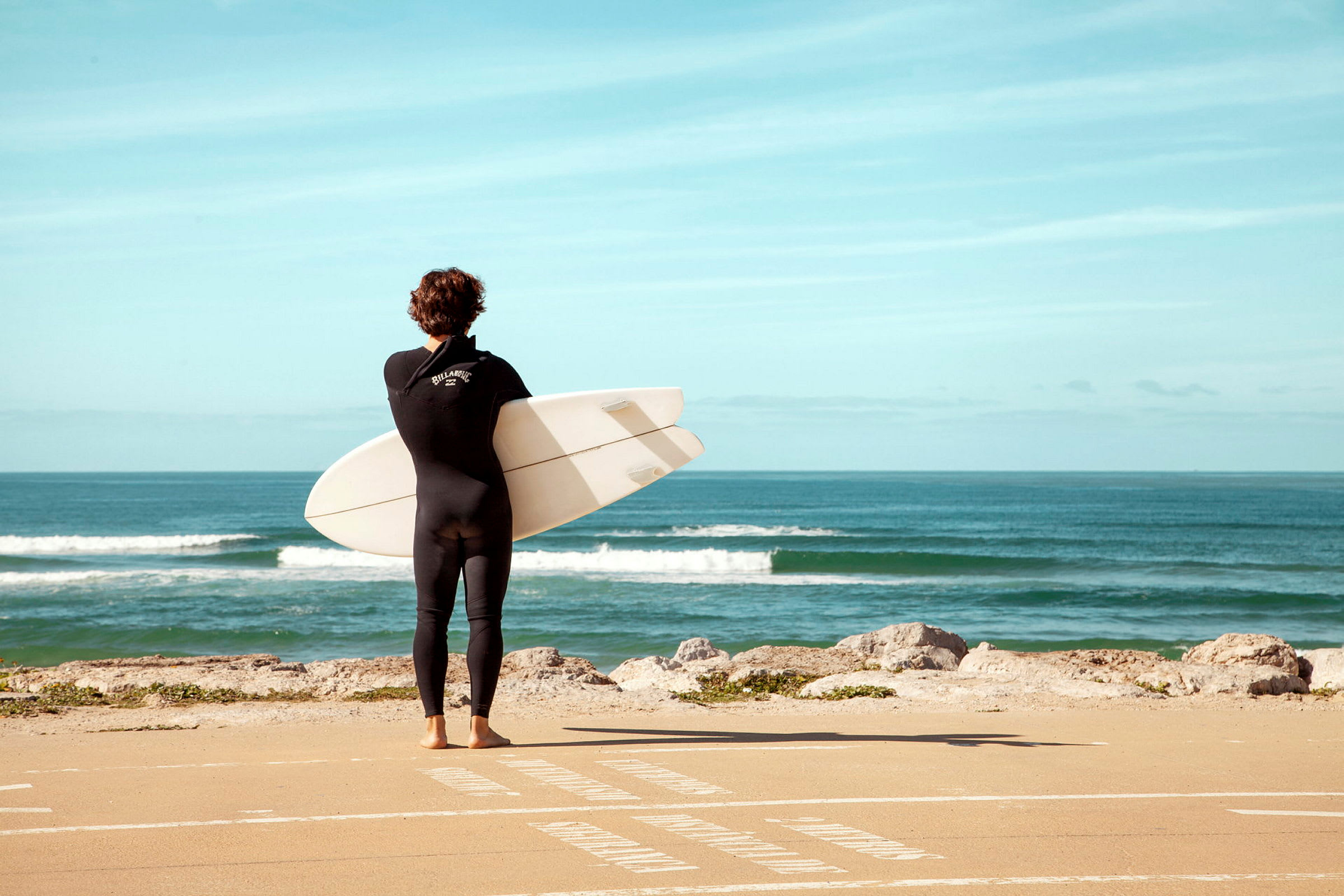 En person i våtdräkt håller en surfbräda och står vänd mot havet. (AI-genererad text)