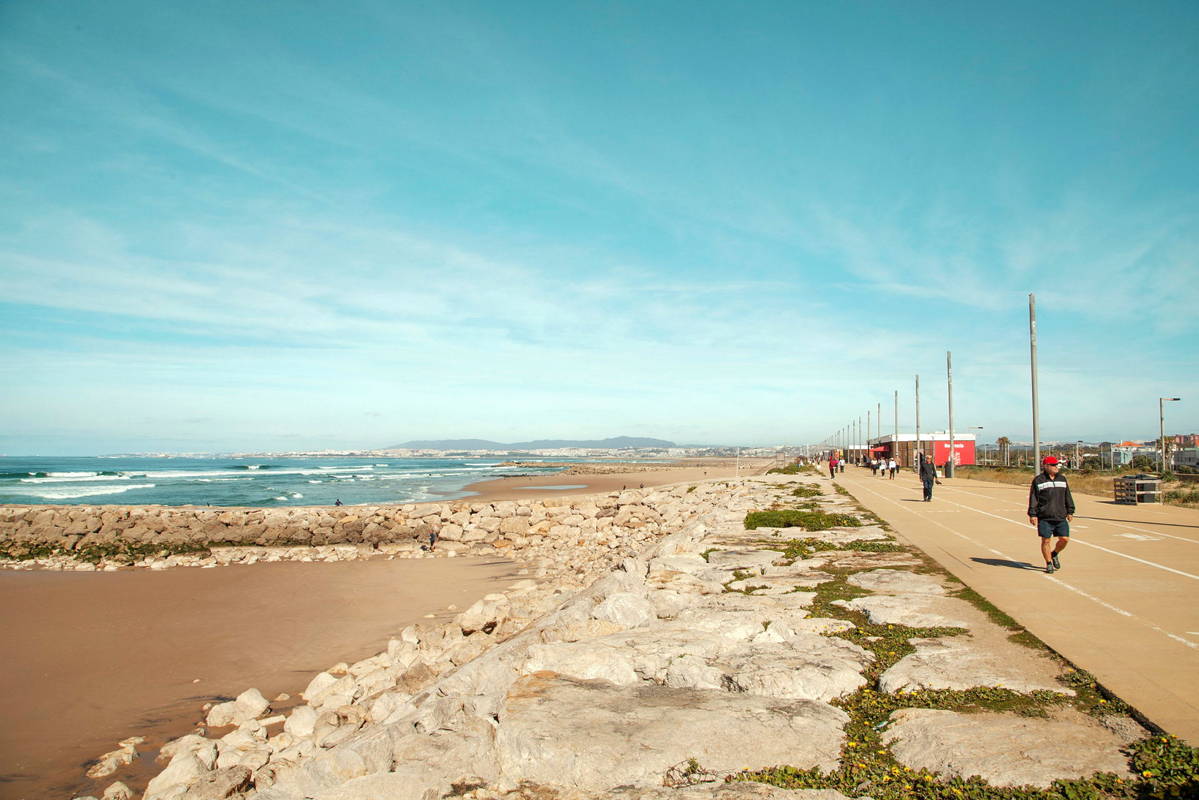 En strandpromenad med en gångväg bredvid havet och klippor, människor promenerar. (AI-genererad text)