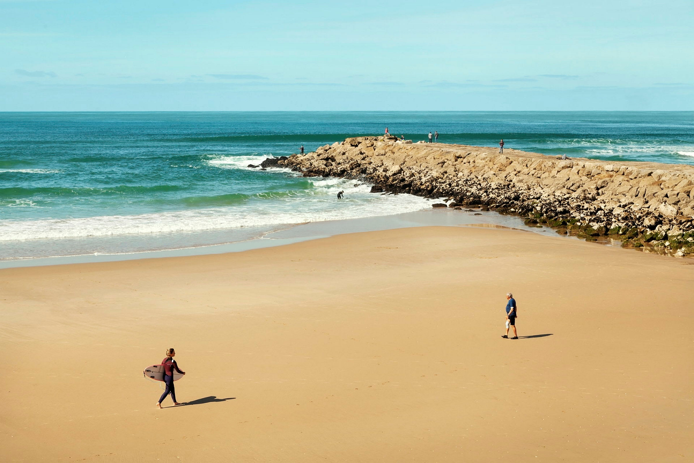 En strand med några personer, en av dem bär en surfbräda. (AI-genererad text)