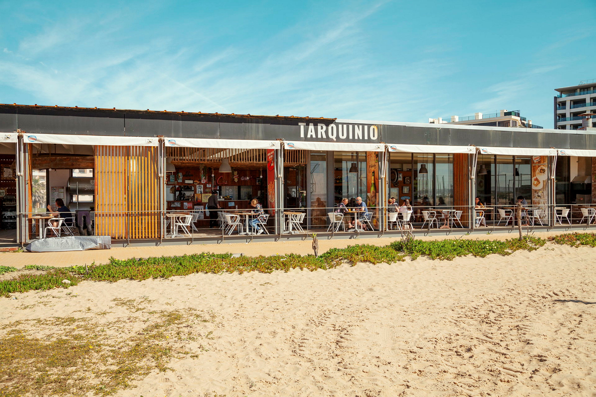 En strandrestaurang med skylt som säger 'Tarquinio', sittplatser utomhus och sandstrand i förgrunden. (AI-genererad text)