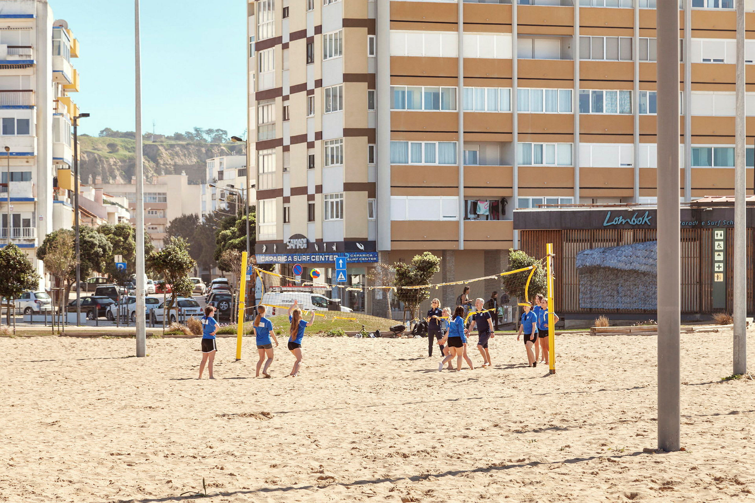 En grupp människor spelar volleyboll på en sandplan med byggnader i bakgrunden. (AI-genererad text)
