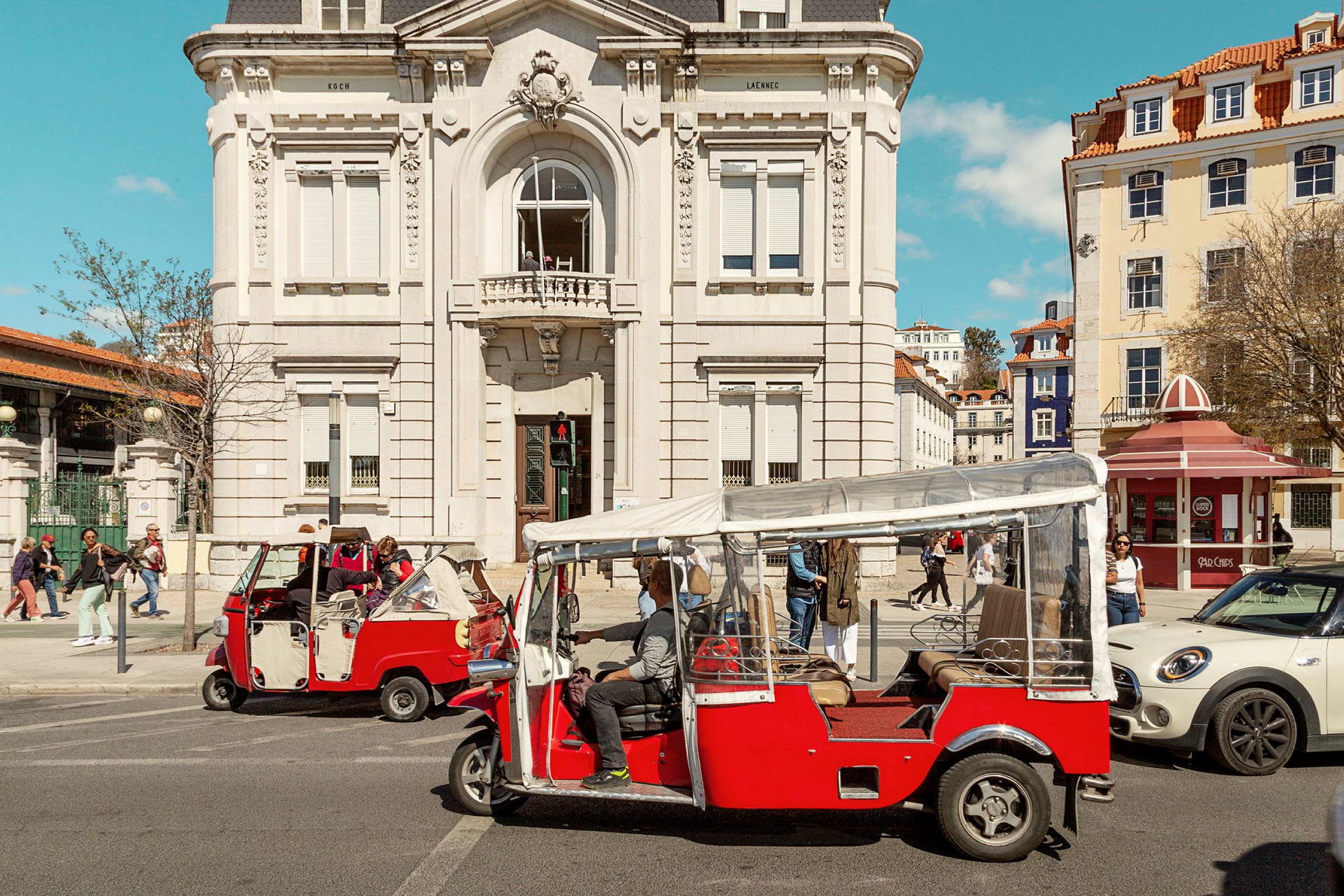 Röda tuk-tuk-fordon framför en klassisk byggnad. (AI-genererad text)