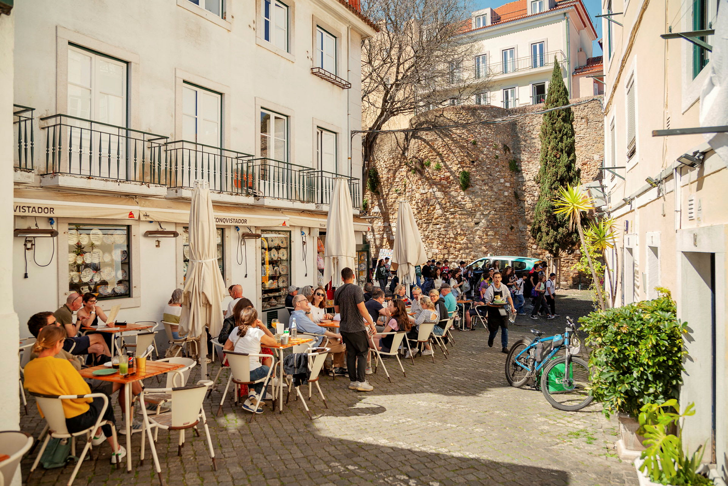 En utomhusterrass vid en restaurang med flera människor som sitter vid bord och njuter av sin måltid. I bakgrunden syns en stenmur och en trädbeklädd byggnad. (AI-genererad text)