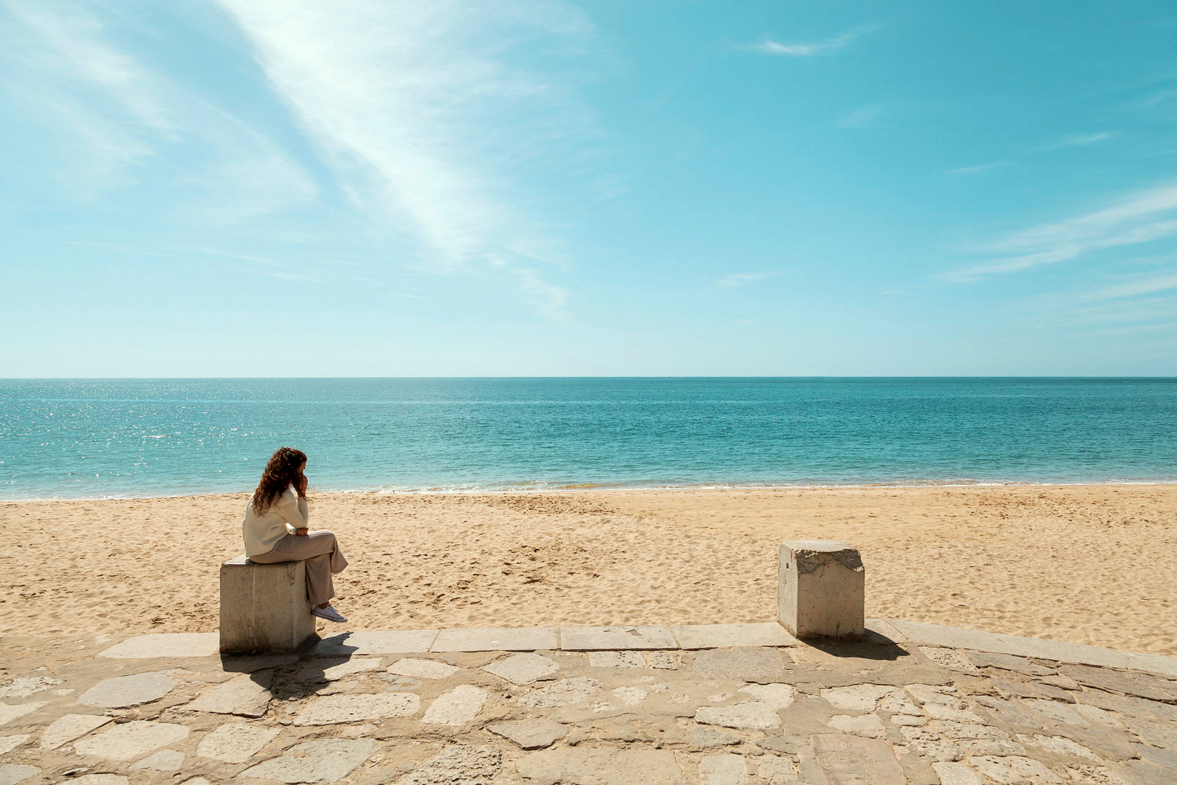 En person sitter på en betongblock vid en sandstrand med utsikt över havet. (AI-genererad text)