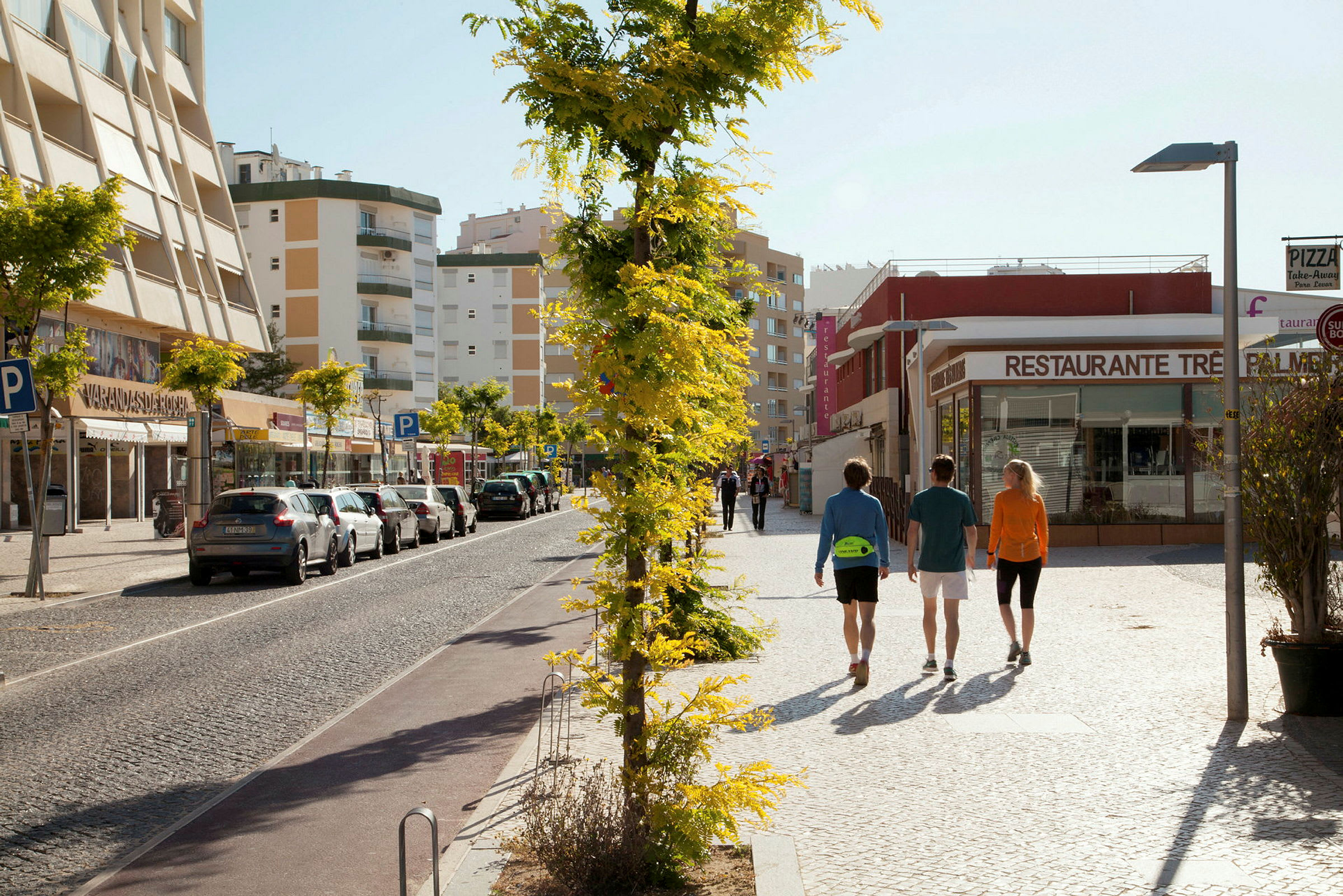Personer promenerar på en gata med parkerade bilar och byggnader i bakgrunden. (AI-genererad text)