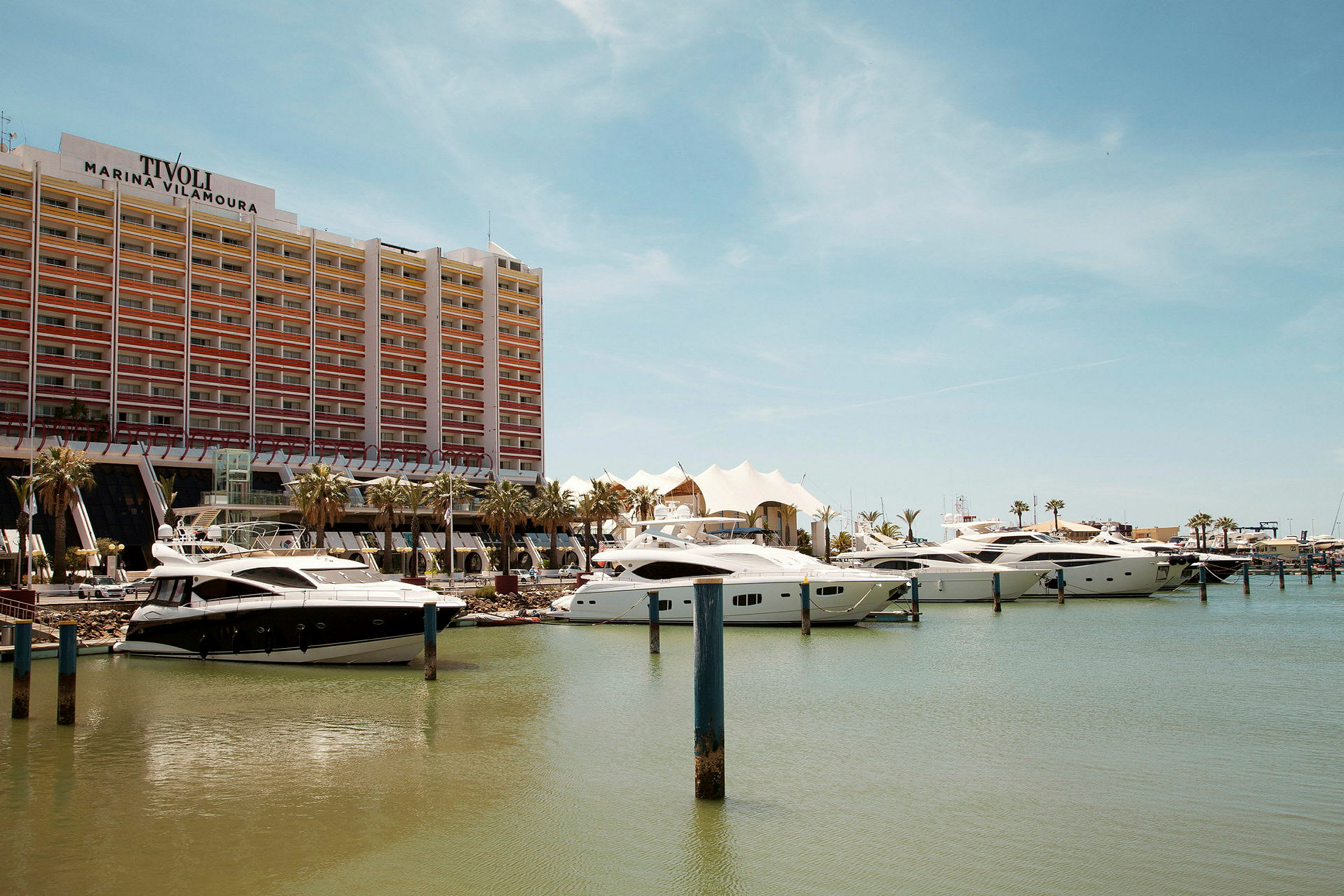 En modern byggnad med Tivoli Marina Vilamoura-logga bakom förtöjda yachter vid en småbåtshamn. (AI-genererad text)