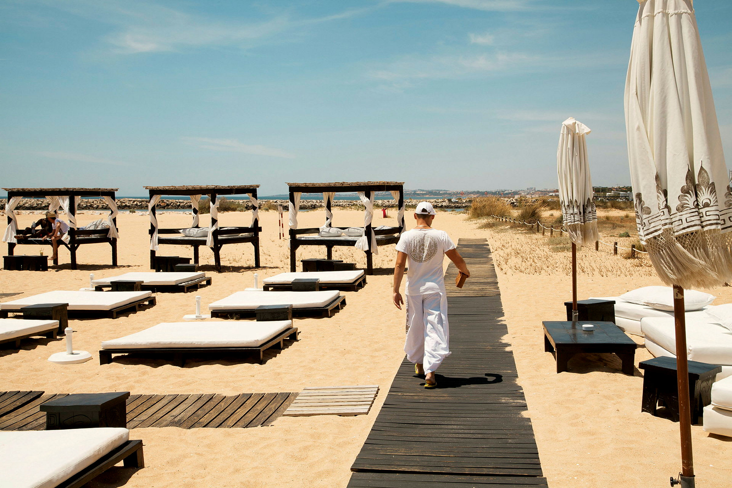 Man promenerar på träbrygga vid strand med solstolar och solbäddar. (AI-genererad text)