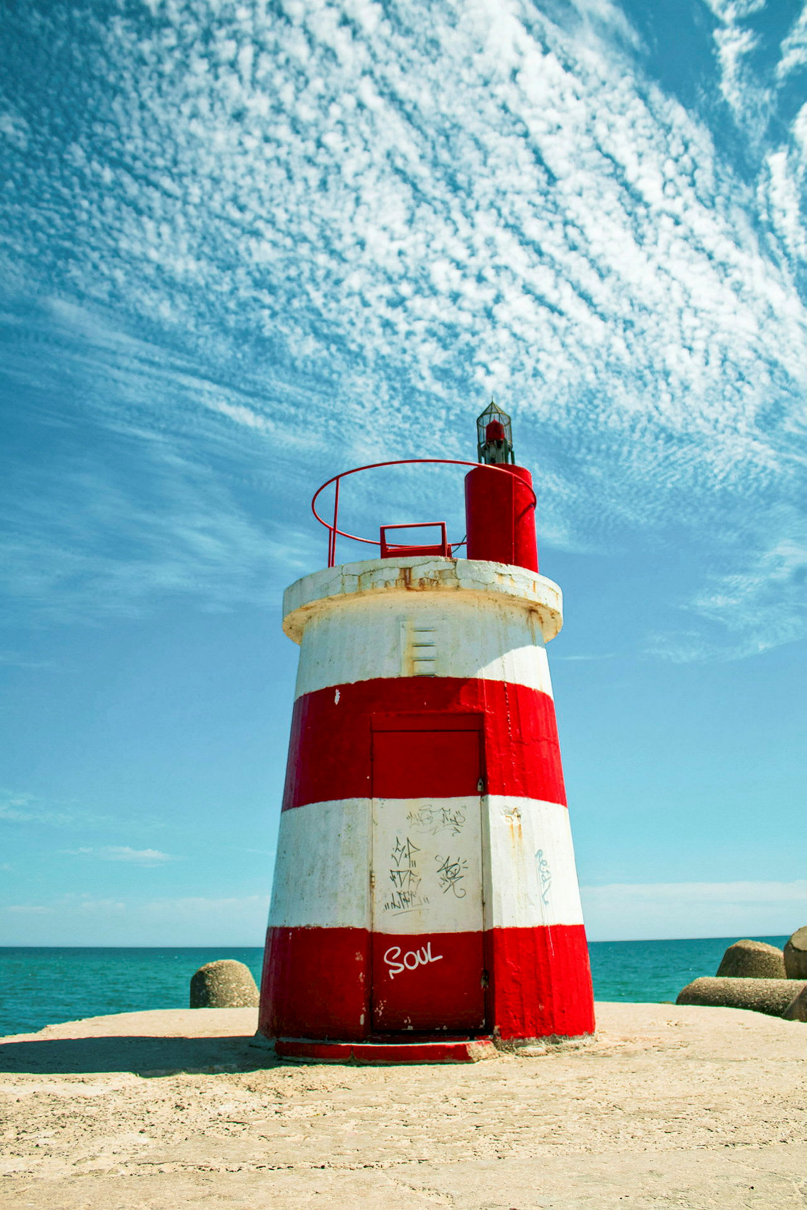 En fyren med röda och vita horisontella ränder står på en betonggrund vid havet med en blå himmel i bakgrunden. (AI-genererad text)