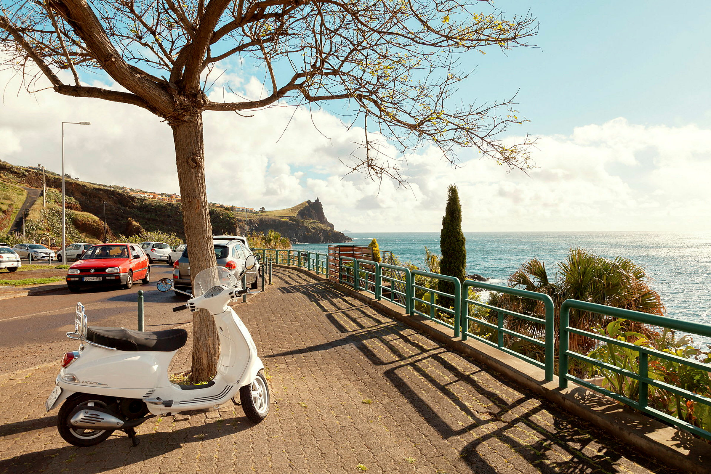 En vit moped står parkerad vid en trottoar nära havet. Träd och bilar syns i bakgrunden. (AI-genererad text)