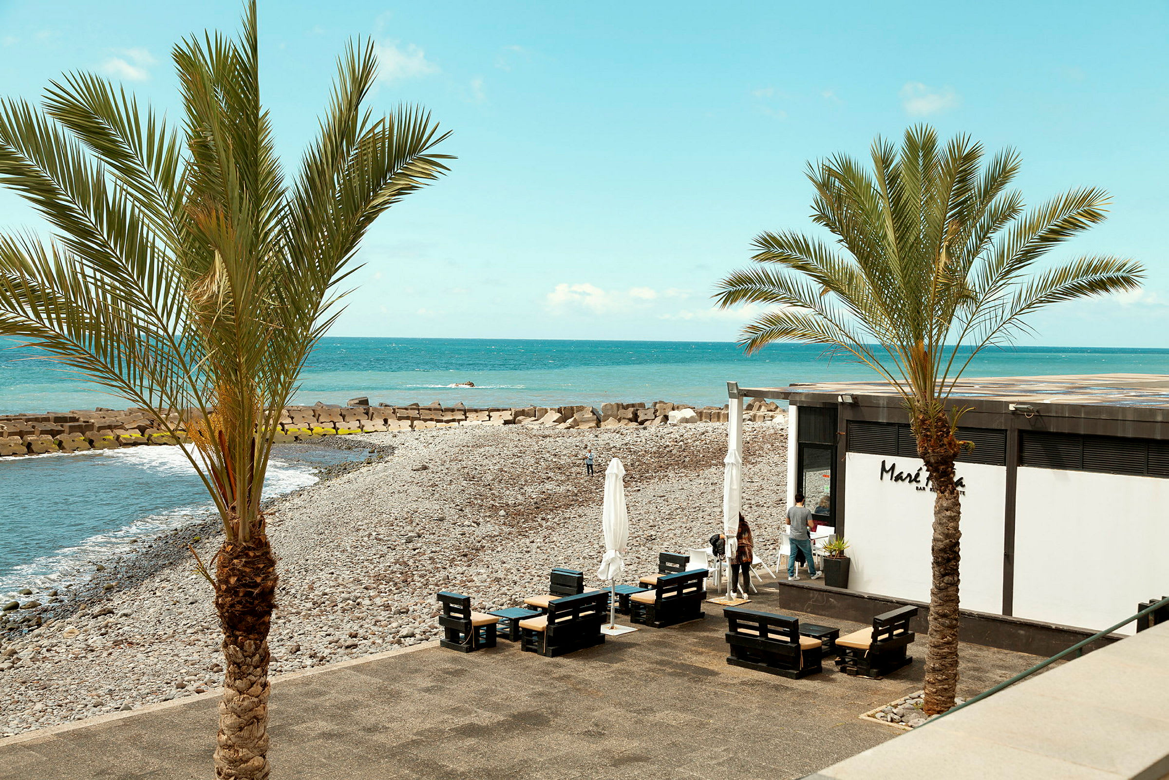 En stenstrand med palmer och ett kafé vid havet. (AI-genererad text)