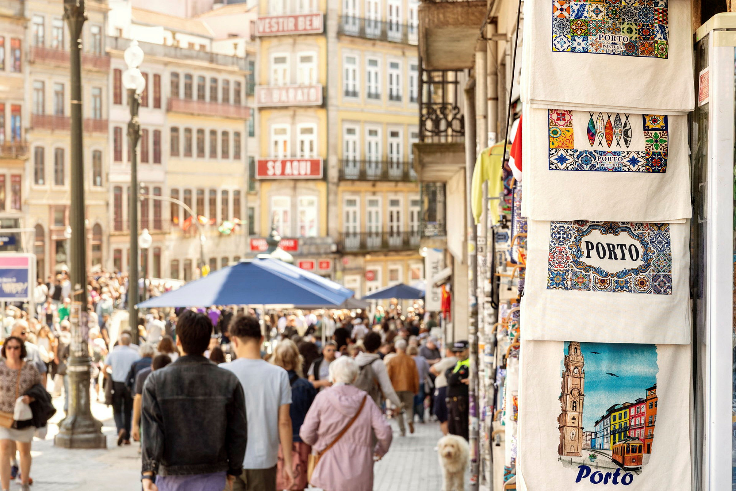 Gatan i Porto med folkmassa och byggnader i bakgrunden samt tygväskor som hänger till försäljning. (AI-genererad text)