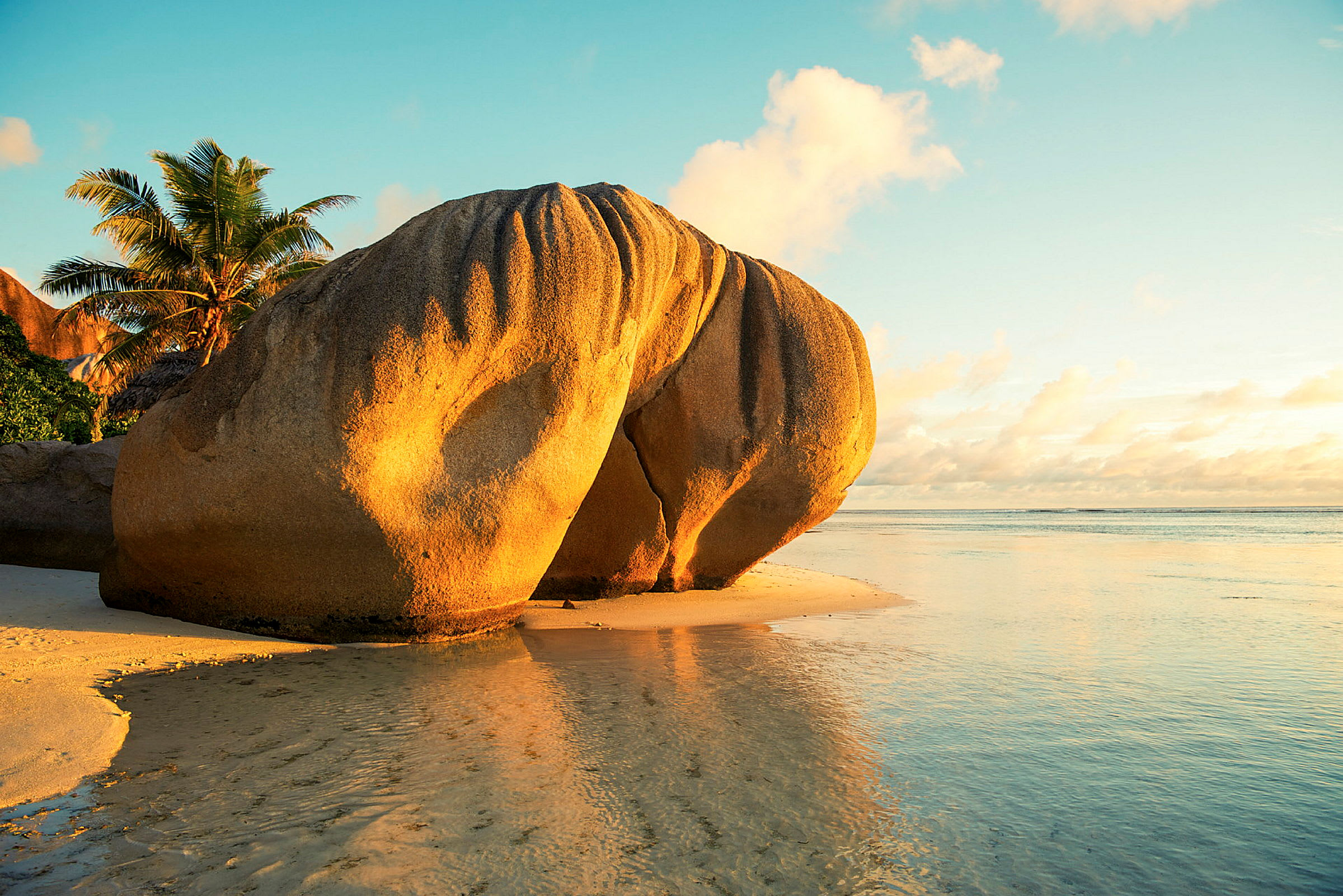 Ett stort sandstensblock står på stranden medan solen går ner, och en palm syns i bakgrunden. (AI-genererad text)