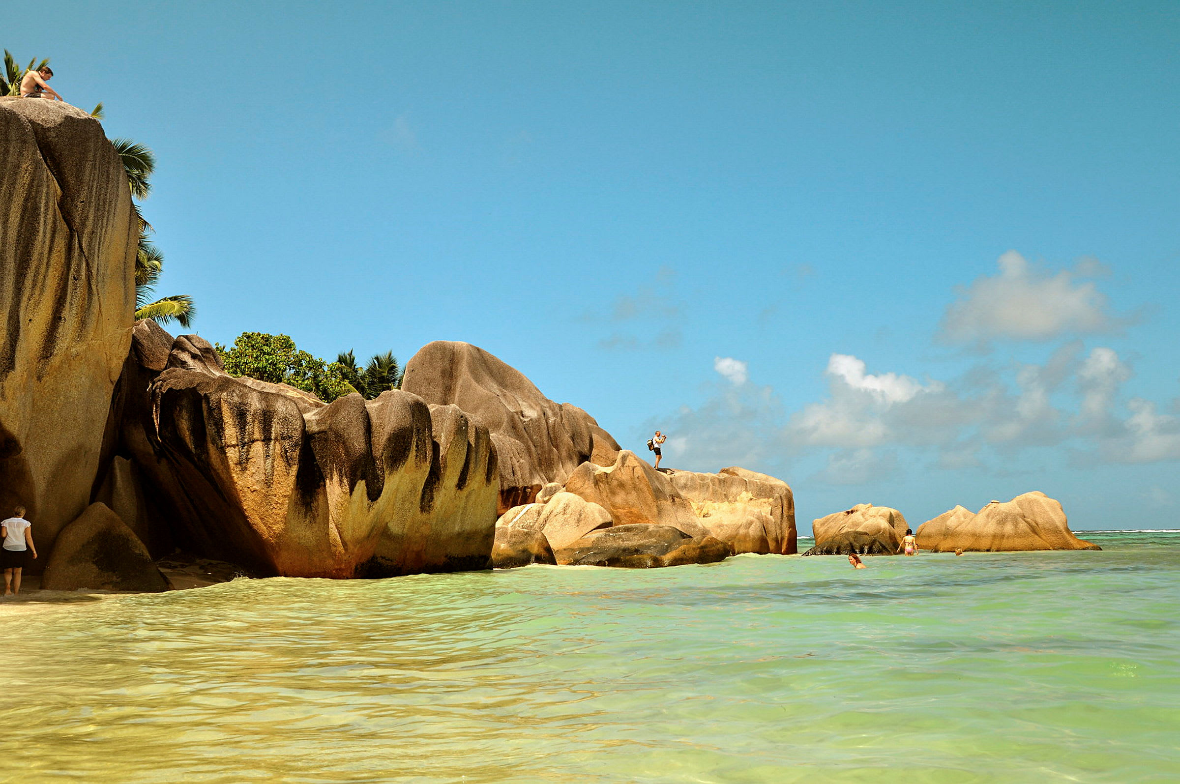 Strand med stora stenformationer omgivna av klart vatten. En person står på en sten och andra syns vid stranden. (AI-genererad text)