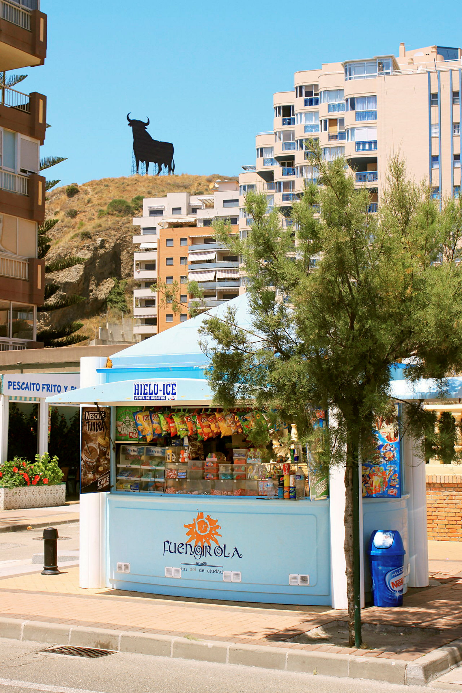 En kiosk omgiven av palmer med bakgrund av byggnader och en tjursiluett på en kulle. (AI-genererad text)