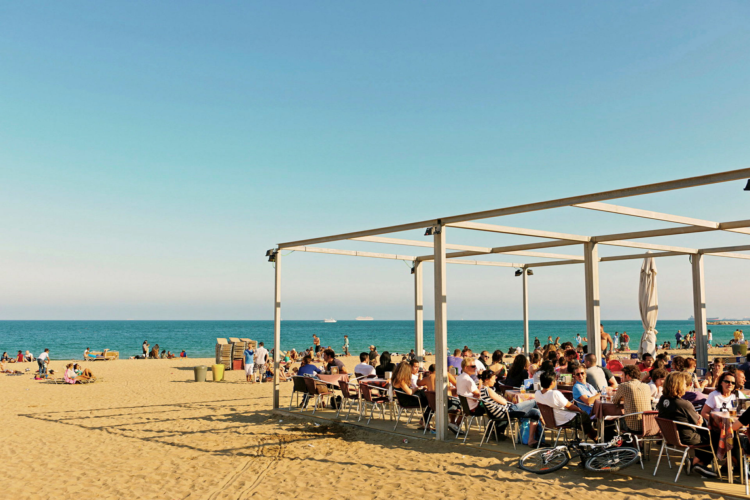 En strand med människor som sitter vid ett caféområde under ett tak och andra som njuter av stranden och havet. (AI-genererad text)