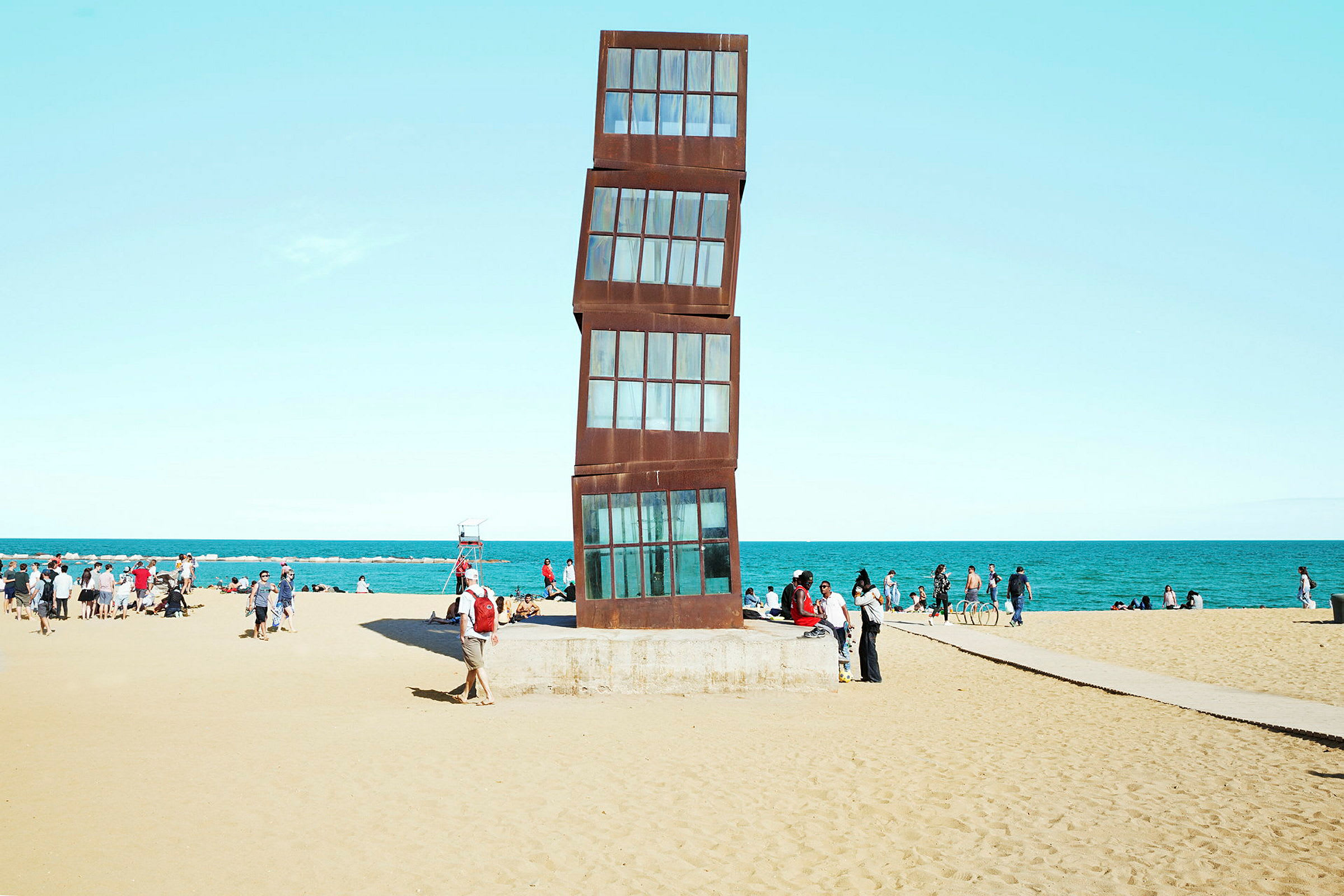 Bild av en strand med en modern konstskulptur i fokus samt människor i bakgrunden. (AI-genererad text)