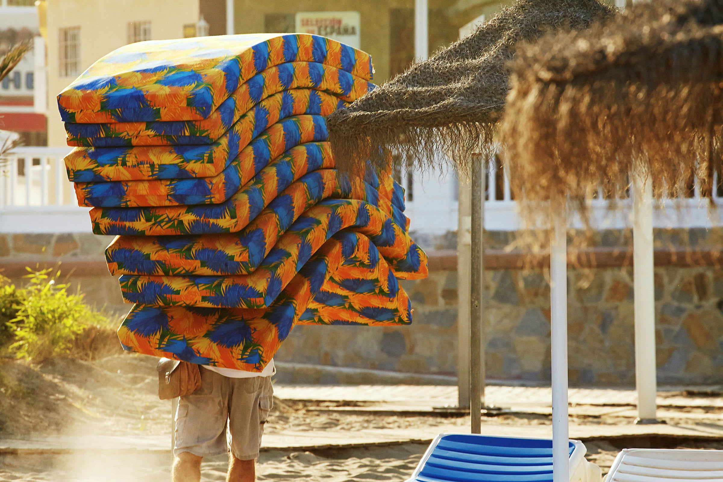 En person bär en stor hög av färgglada madrasser på en strand. (AI-genererad text)