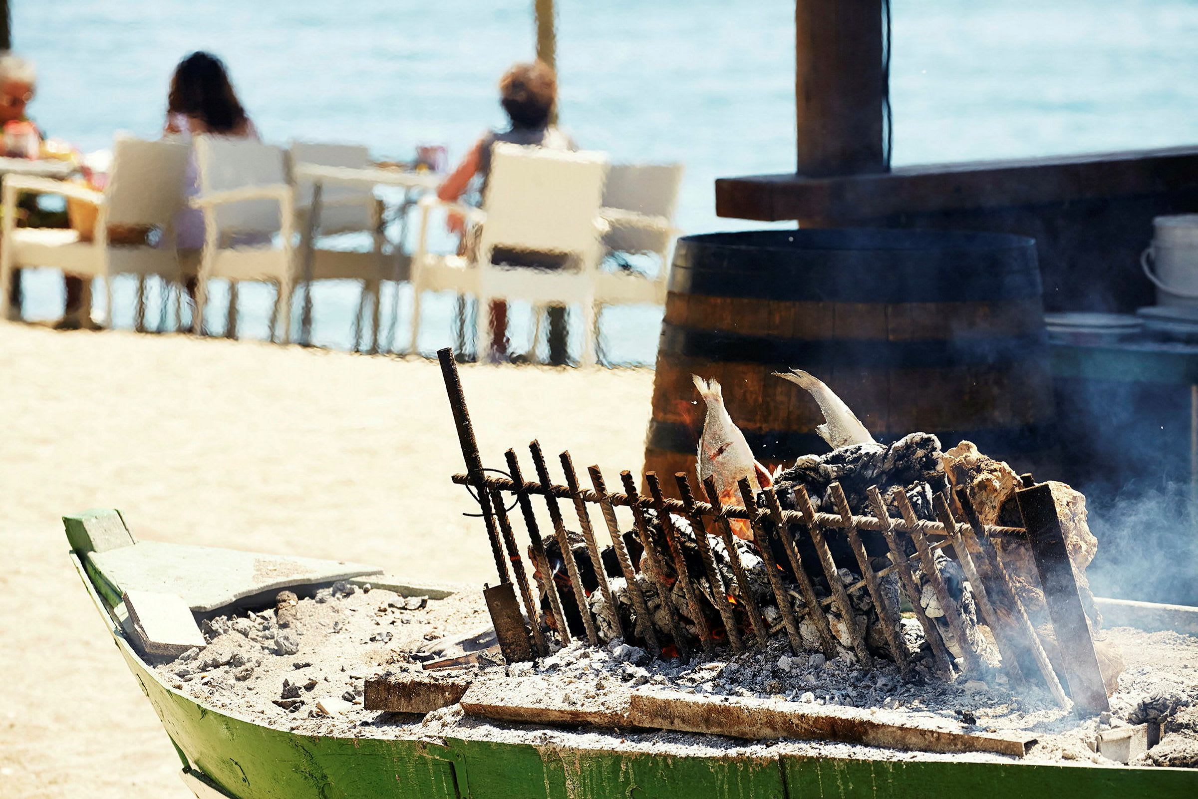 Grillade fiskar på ett spett över glöd, med personer sittande vid strandbord i bakgrunden. (AI-genererad text)