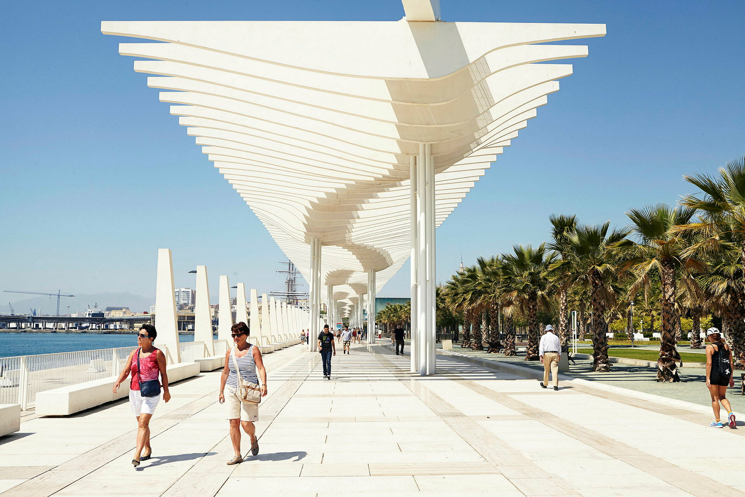 En strandpromenad med modern arkitektur och palmer på en solig dag. Människor promenerar längs gångvägen. (AI-genererad text)