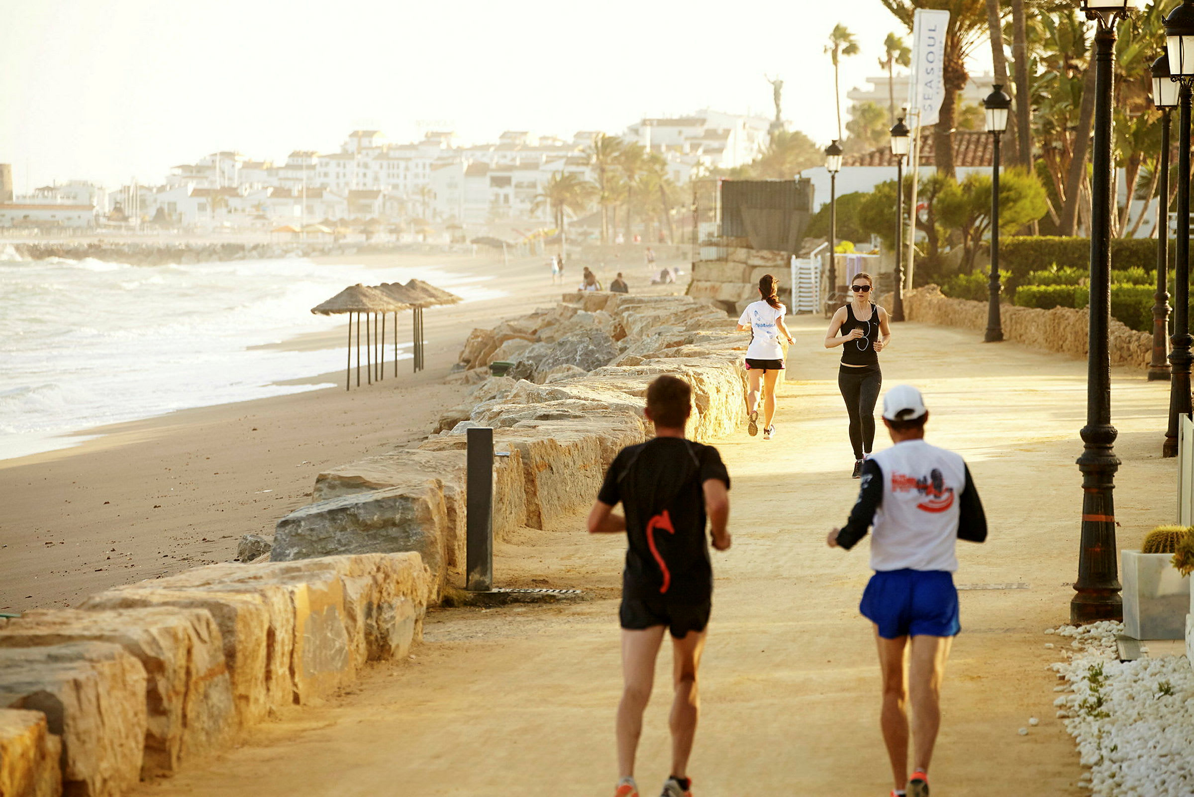 Personer joggar på en strandpromenad nära havet med hus och palmer i bakgrunden. (AI-genererad text)