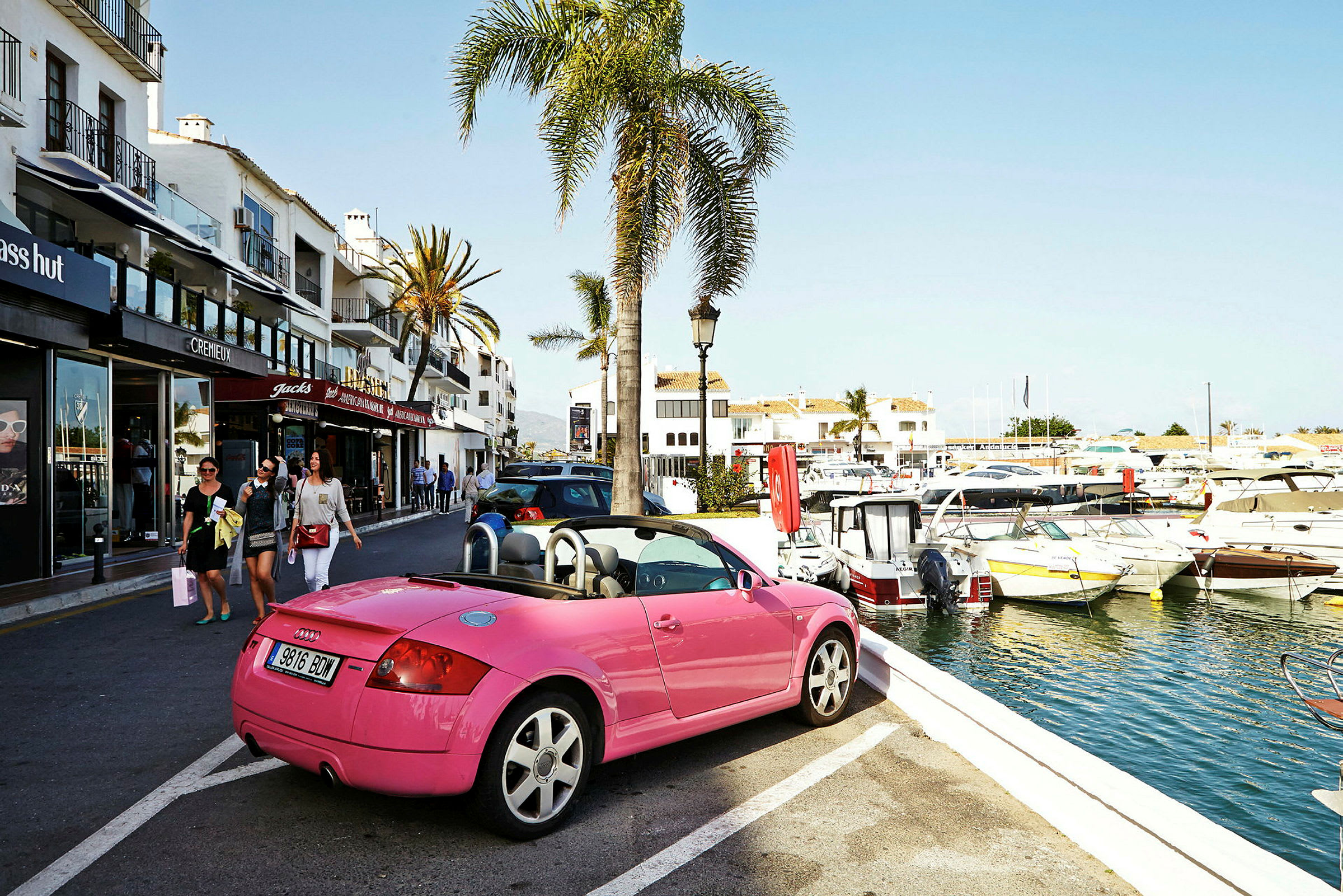 En rosa cabriolet parkerad nära en hamn med båtar, intill en gata med butiker och palmer. (AI-genererad text)