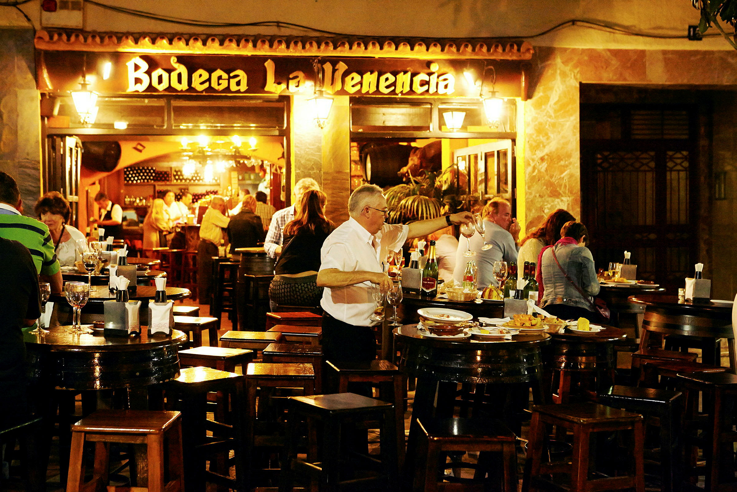 En uteservering med bord och stolar där människor äter och dricker. Ett skylt med texten 'Bodega La Venencia' syns i bakgrunden. (AI-genererad text)