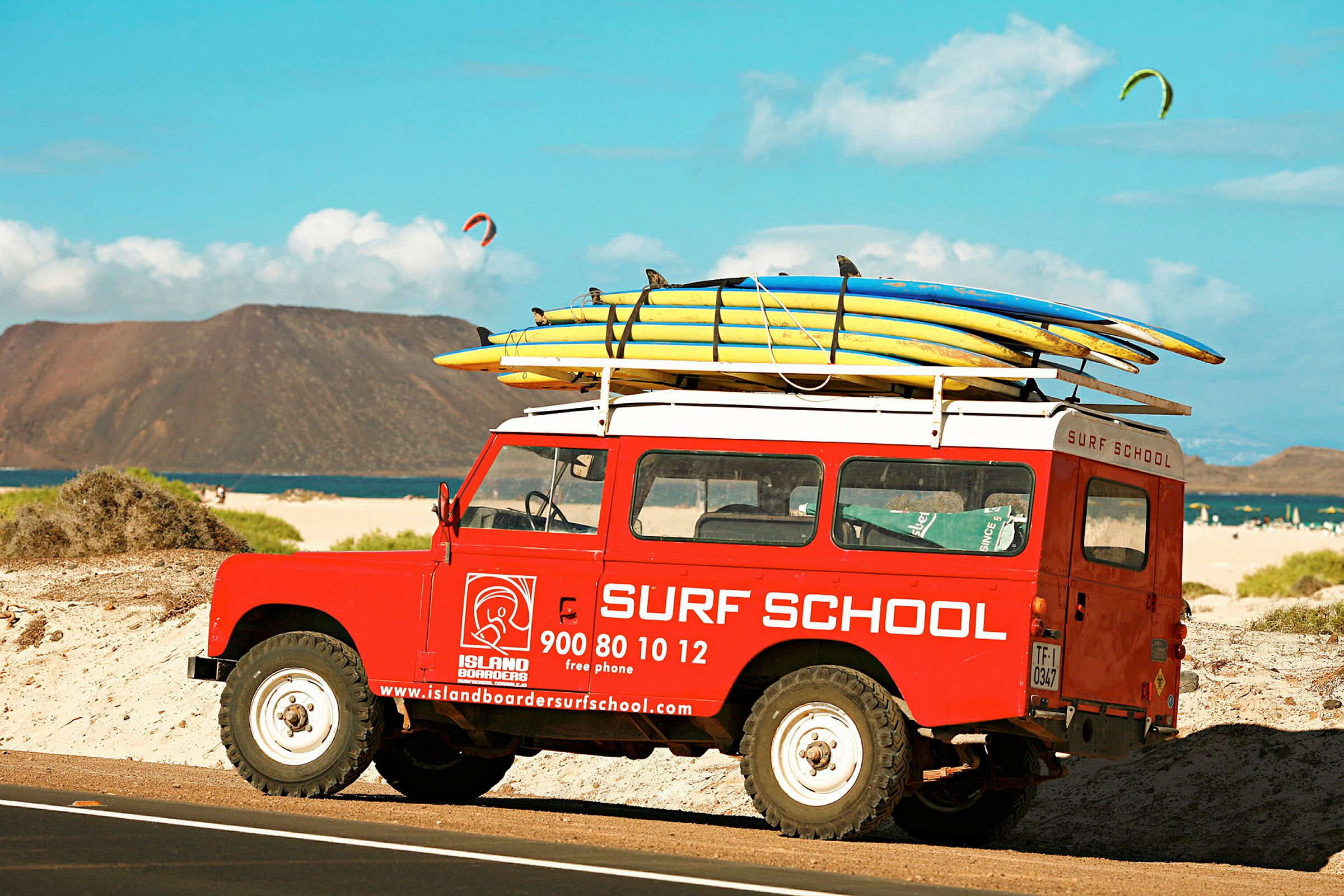En röd bil märkt med "Surf School" står parkerad på en strand med surfbrädor fastspända på taket. (AI-genererad text)