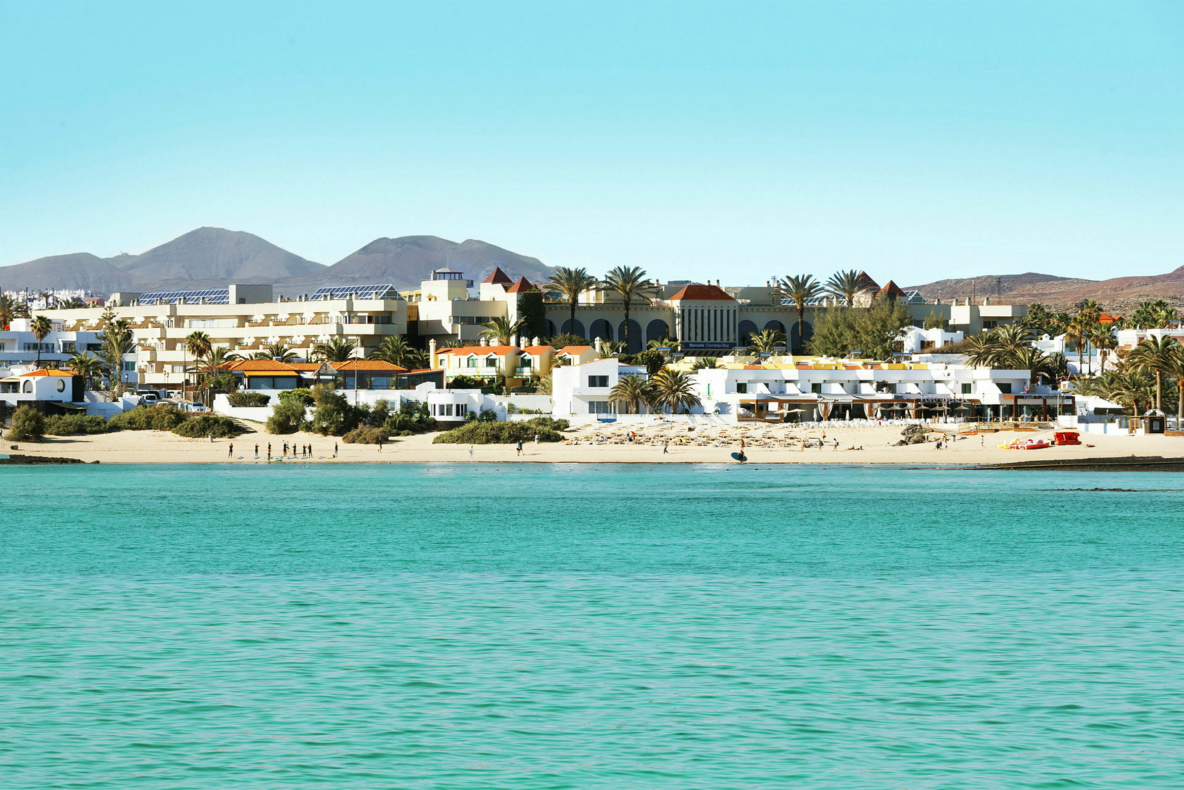 En strand med vit sand och turkost vatten framför en rad av byggnader och palmer. (AI-genererad text)