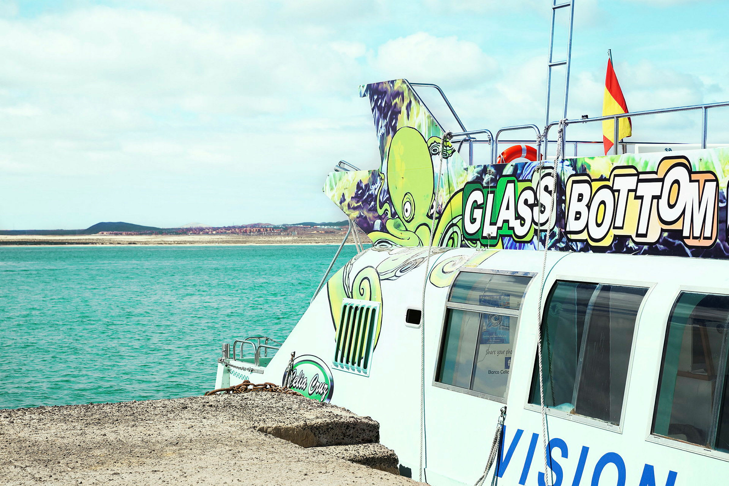 Bild av en båt med glasbotten som ligger vid en kaj intill ett turkost hav. Fartyget har färgglada illustrationer och texten "Glass Bottom" syns tydligt. (AI-genererad text)