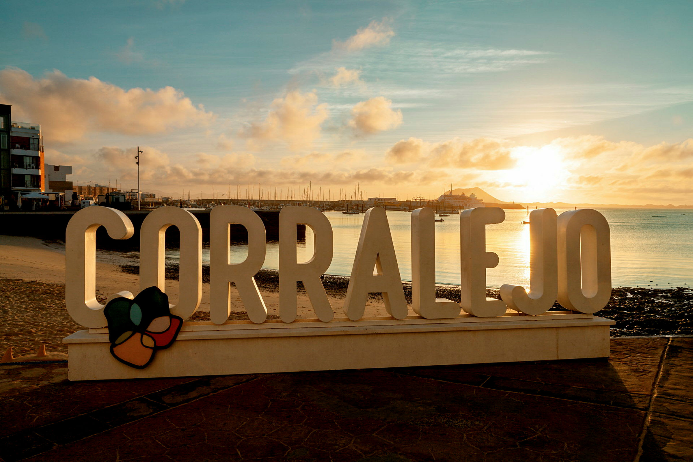 Skylten 'Corralejo' vid en promenadväg med havet och en kaj i bakgrunden under en solnedgång. (AI-genererad text)