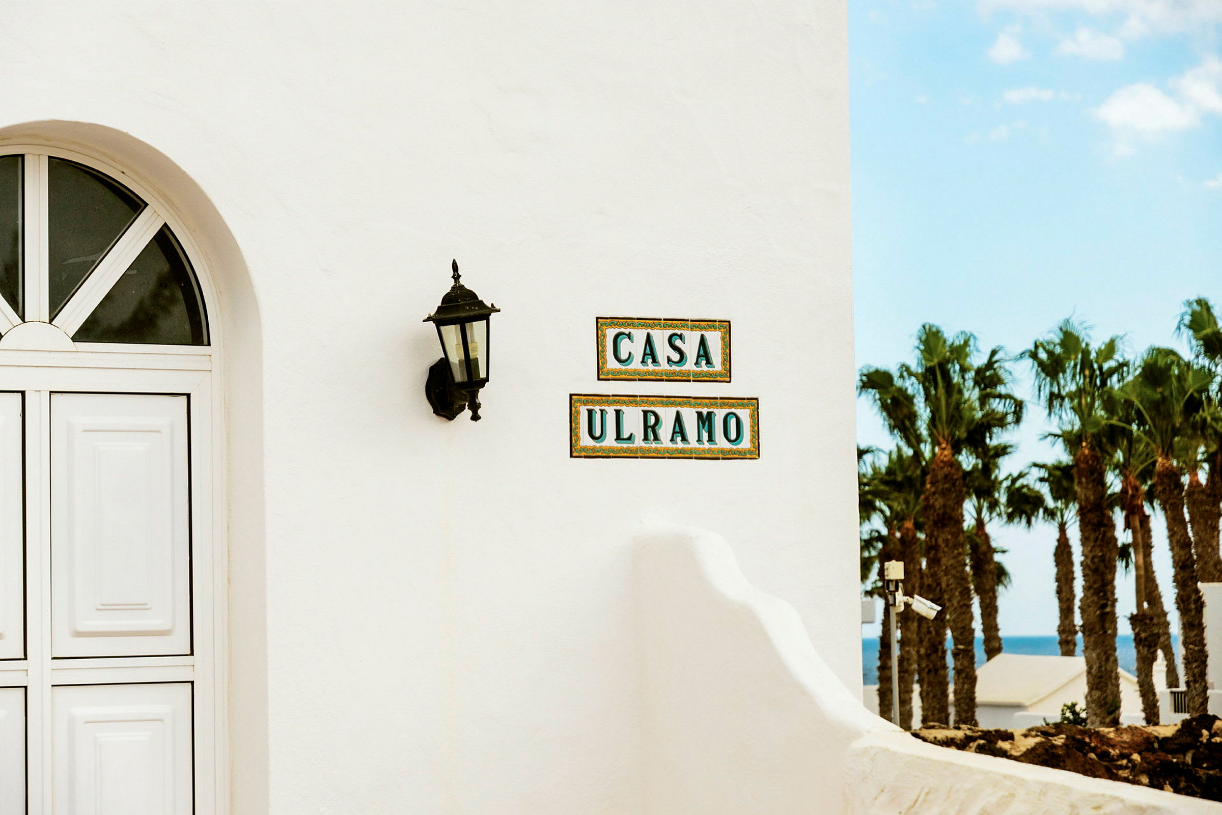 Närbild av en vit byggnadsvägg med ett valvformat fönster, en svart utomhuslykta och plattor med texten 'CASA ULRAMO'. Utsikt över palmer och havet syns i bakgrunden. (AI-genererad text)