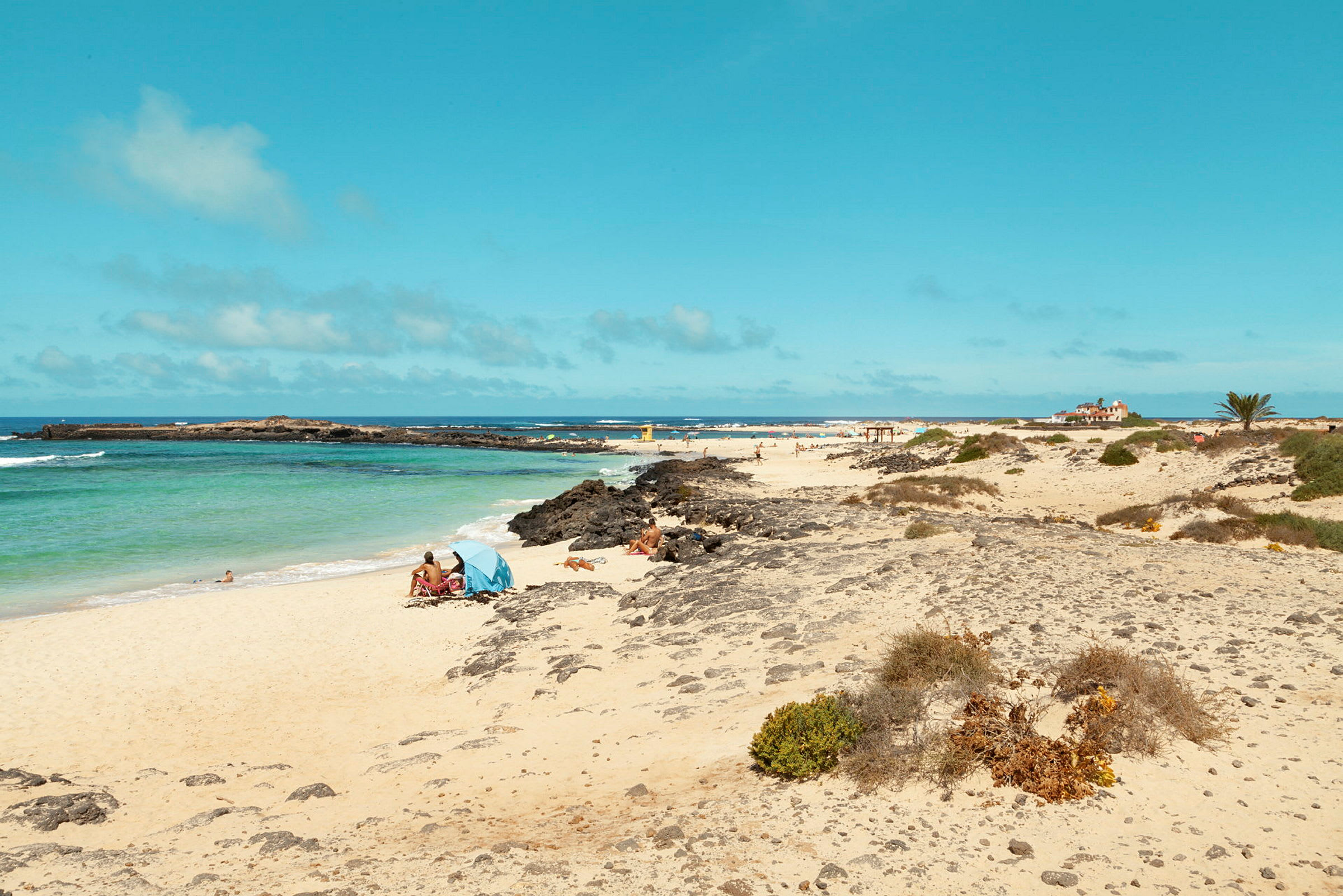 En sandstrand med klippa och växtlighet, samt människor som solar och badar vid det klara vattnet. (AI-genererad text)