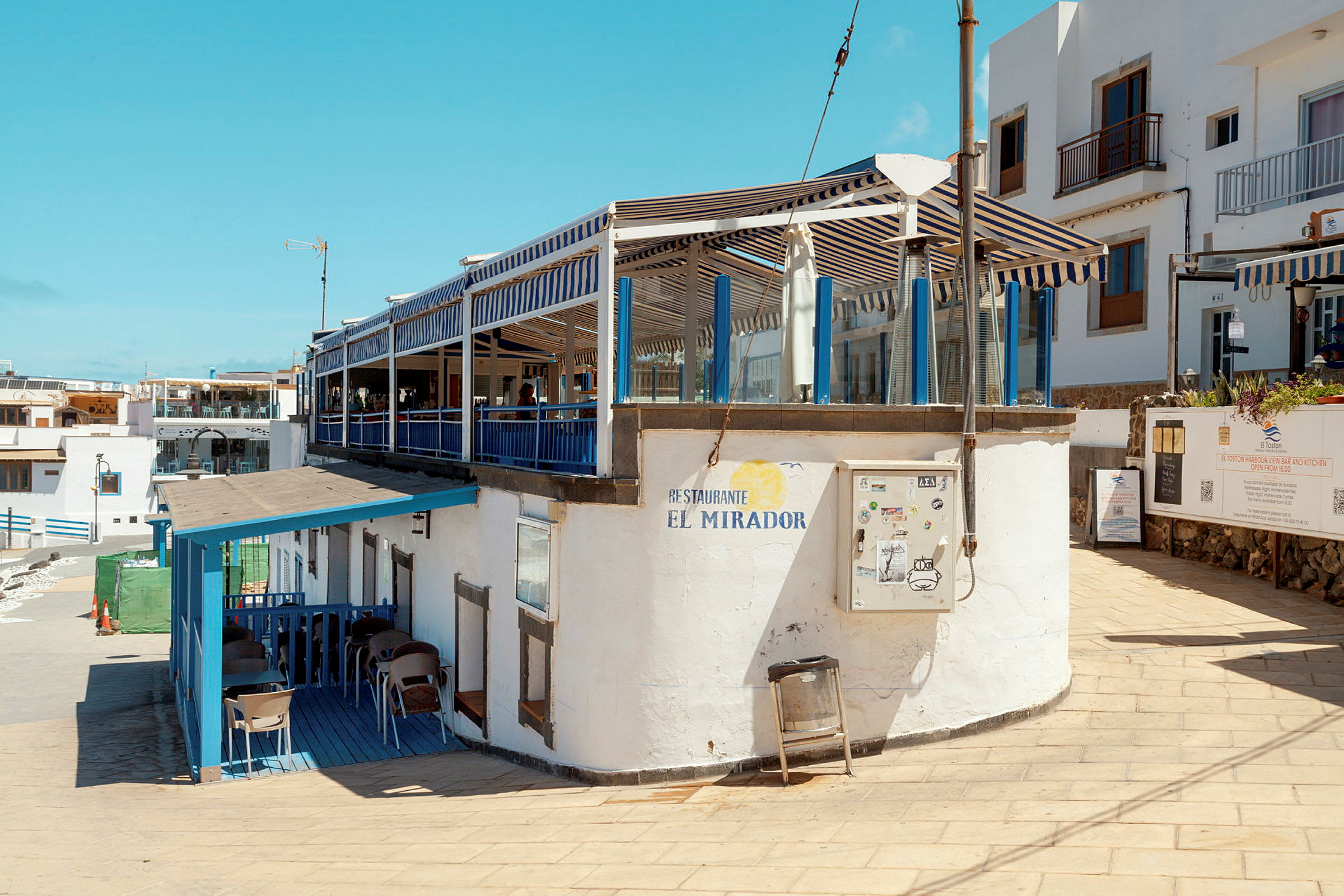 En utomhusrestaurang med blåvitrandiga solskydd och sittplatser under tak, vid en stenlagd stig. (AI-genererad text)