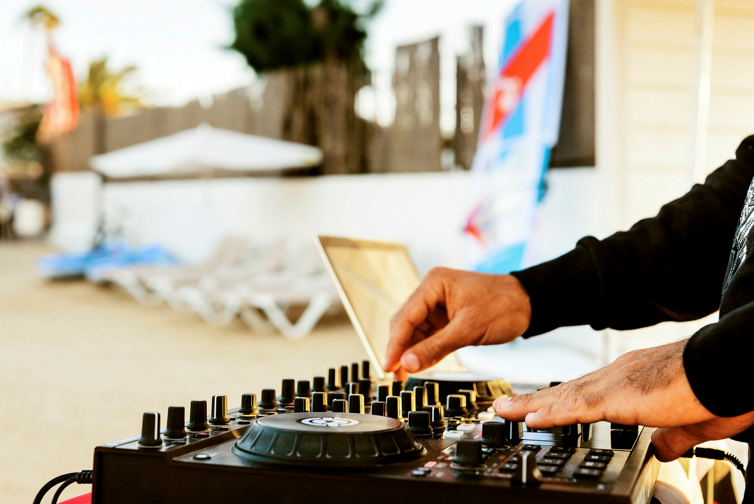 En närbild på en DJ som justerar reglage på en mixer vid en strand med solstolar i bakgrunden. (AI-genererad text)