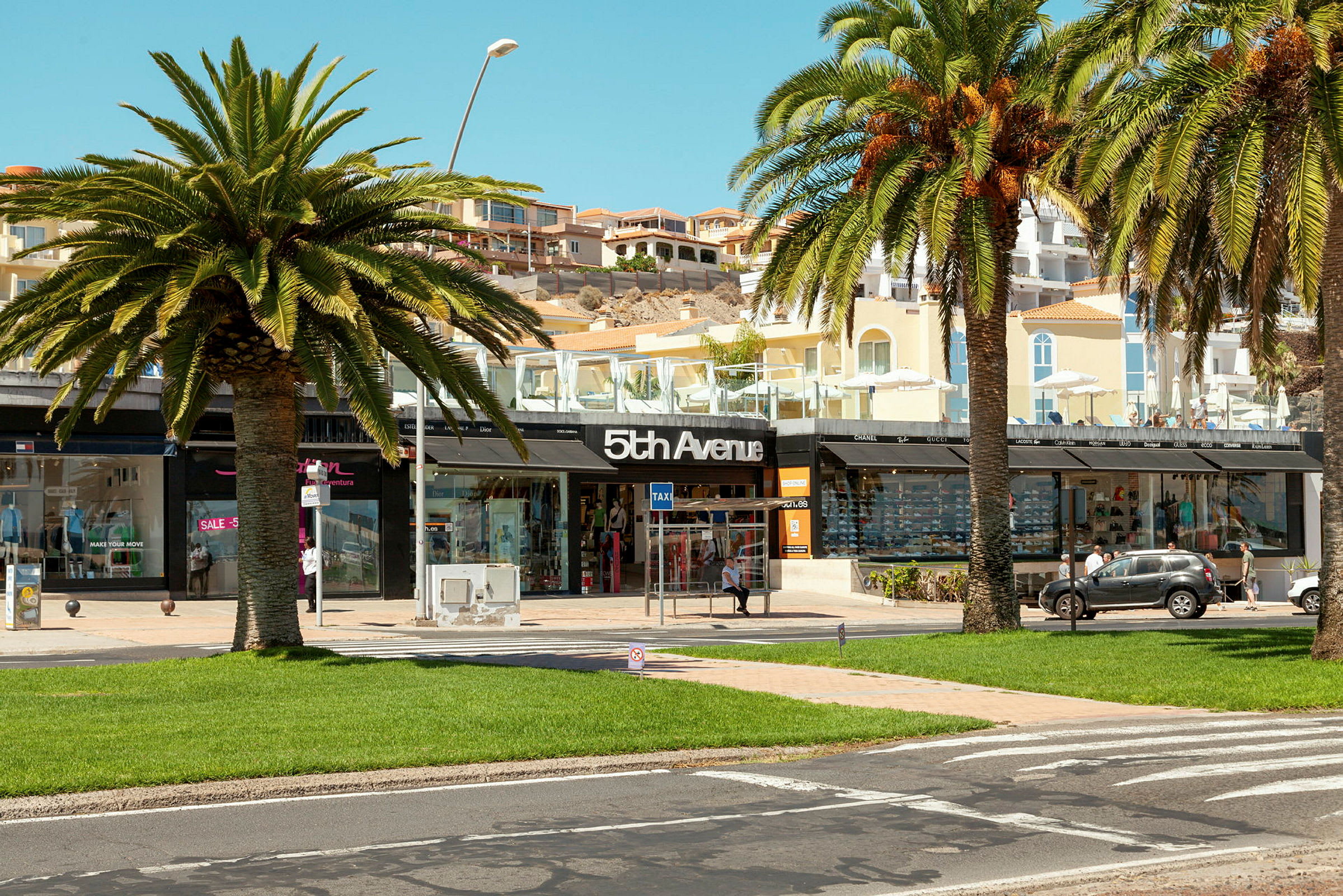 Ett shoppingområde med palmer, grönt gräs och en butik med skylten "5th Avenue". (AI-genererad text)