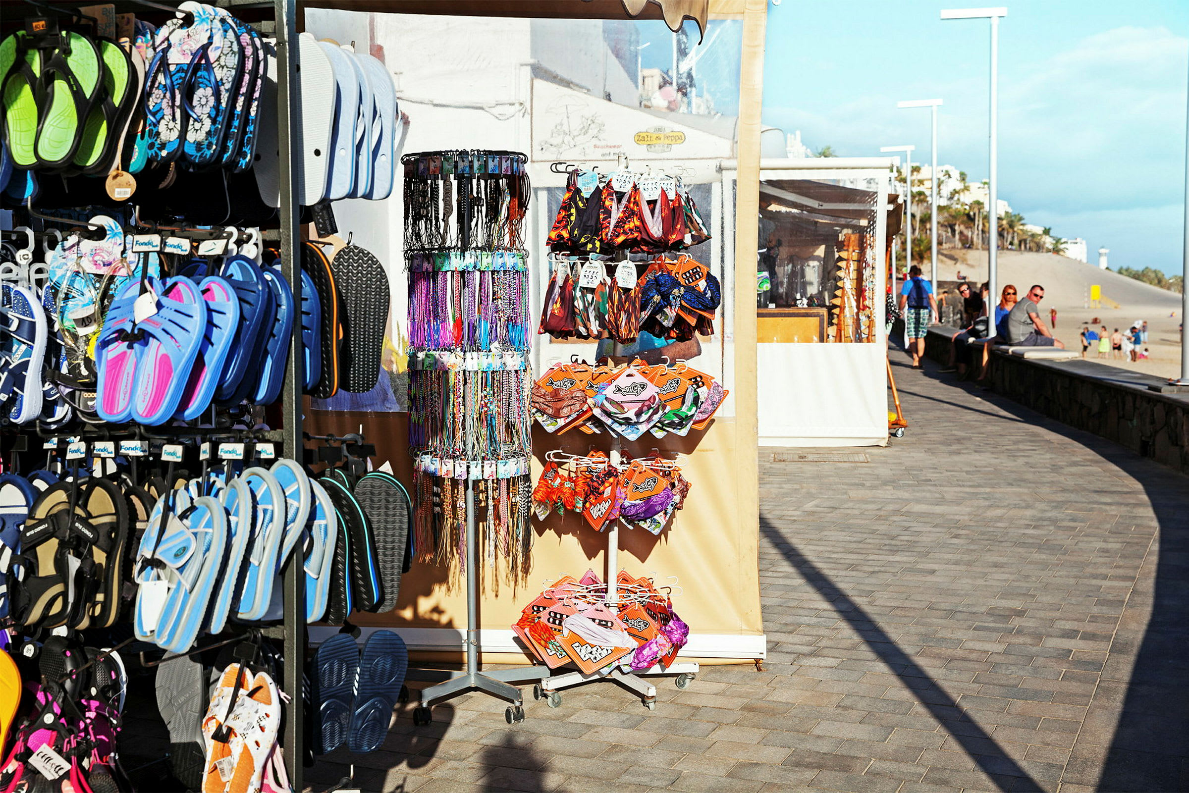 Bild av ett marknadsstånd vid en strandpromenad som säljer flip-flop skor, smycken, och små tygprodukter. (AI-genererad text)