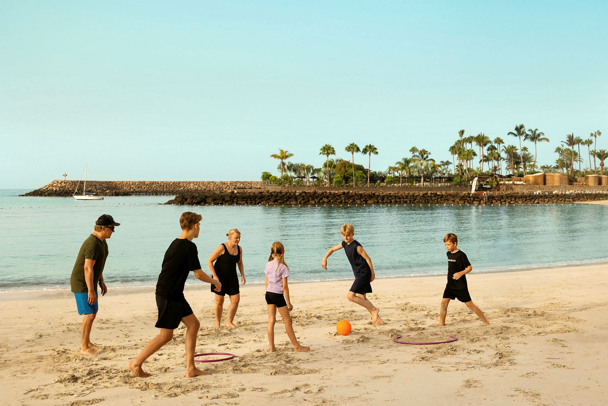 En grupp människor leker med en boll på en sandstrand, med en hamn och palmer i bakgrunden. (AI-genererad text)