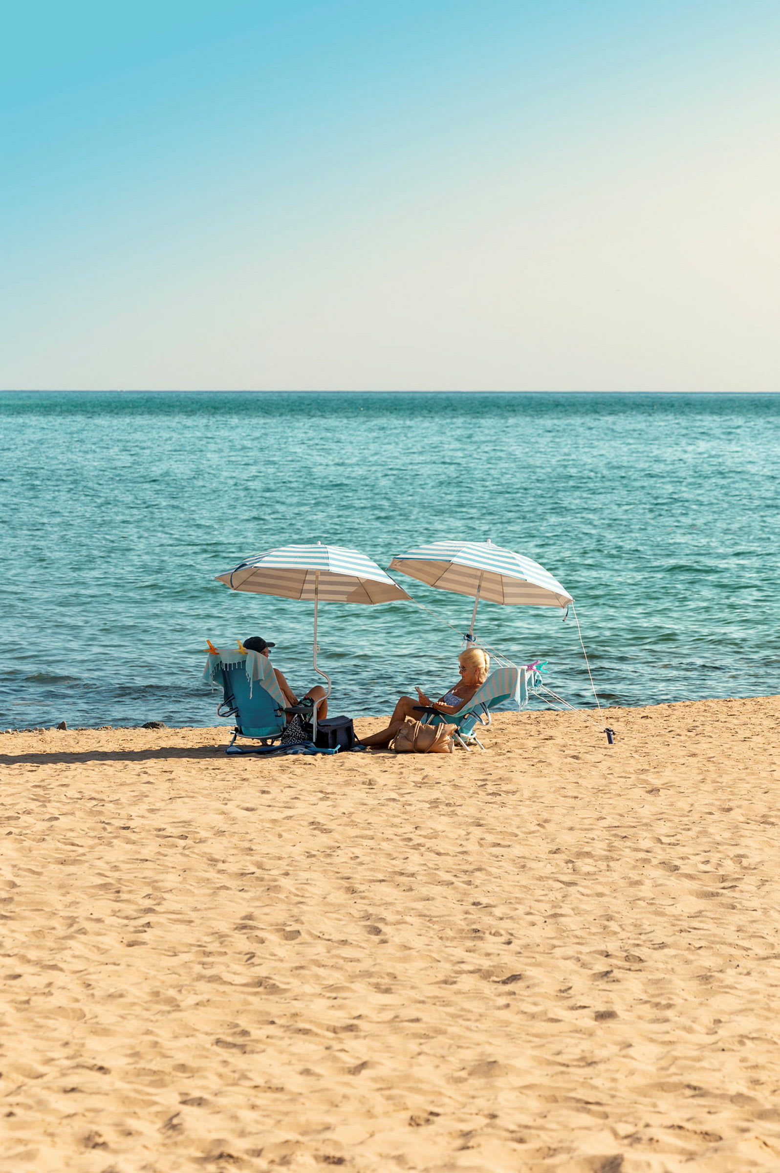 En strand med två personer som sitter under parasoller och njuter av utsikten över havet. (AI-genererad text)