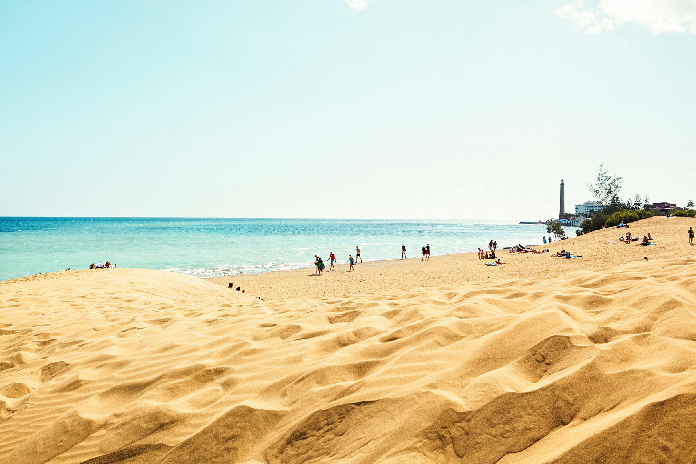 Strand med sanddyner och människor nära havet, med en fyr till höger. (AI-genererad text)