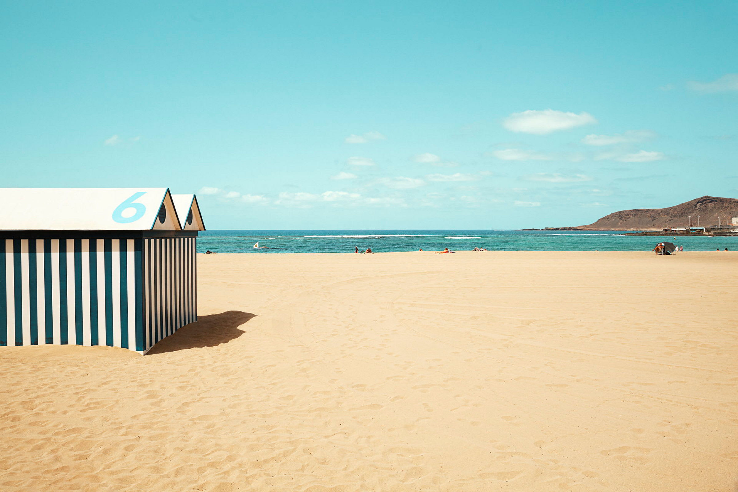 Strand med gul sand, blått hav och himmel, samt ett randigt strandhus med nummer 6 i förgrunden. (AI-genererad text)